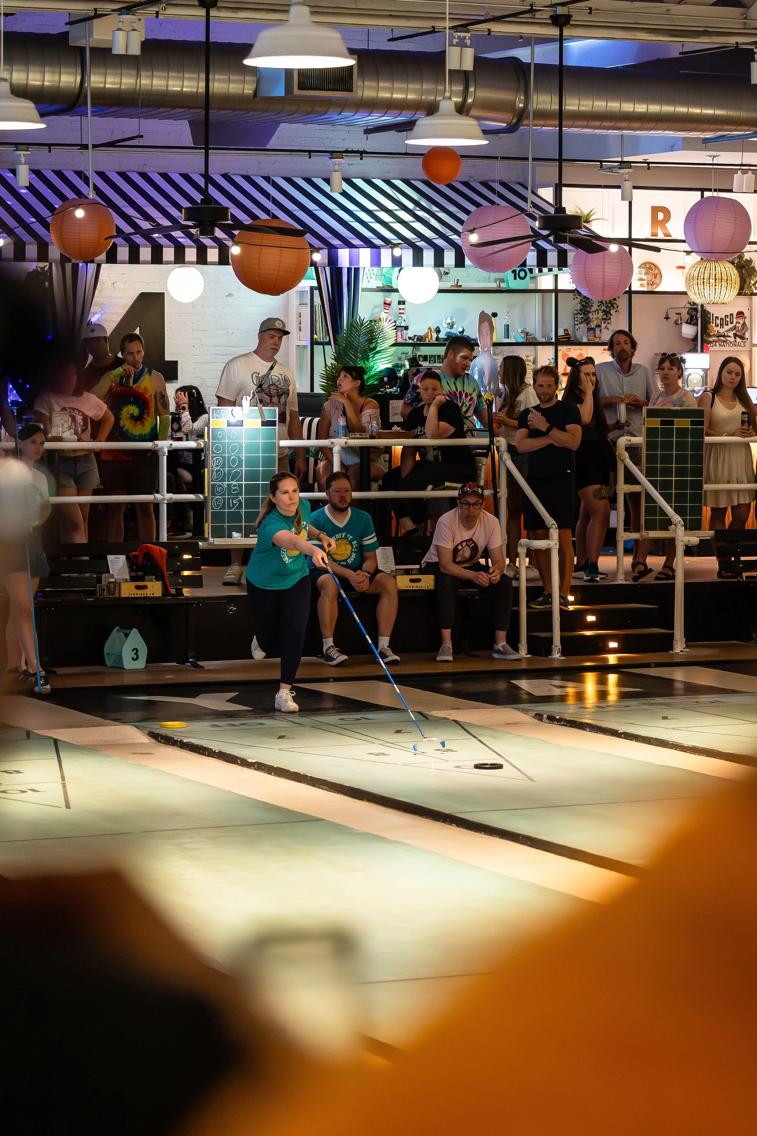 Women playing indoor mini golf at a party or event, with spectators watching from a balcony decorated with hanging paper lanterns