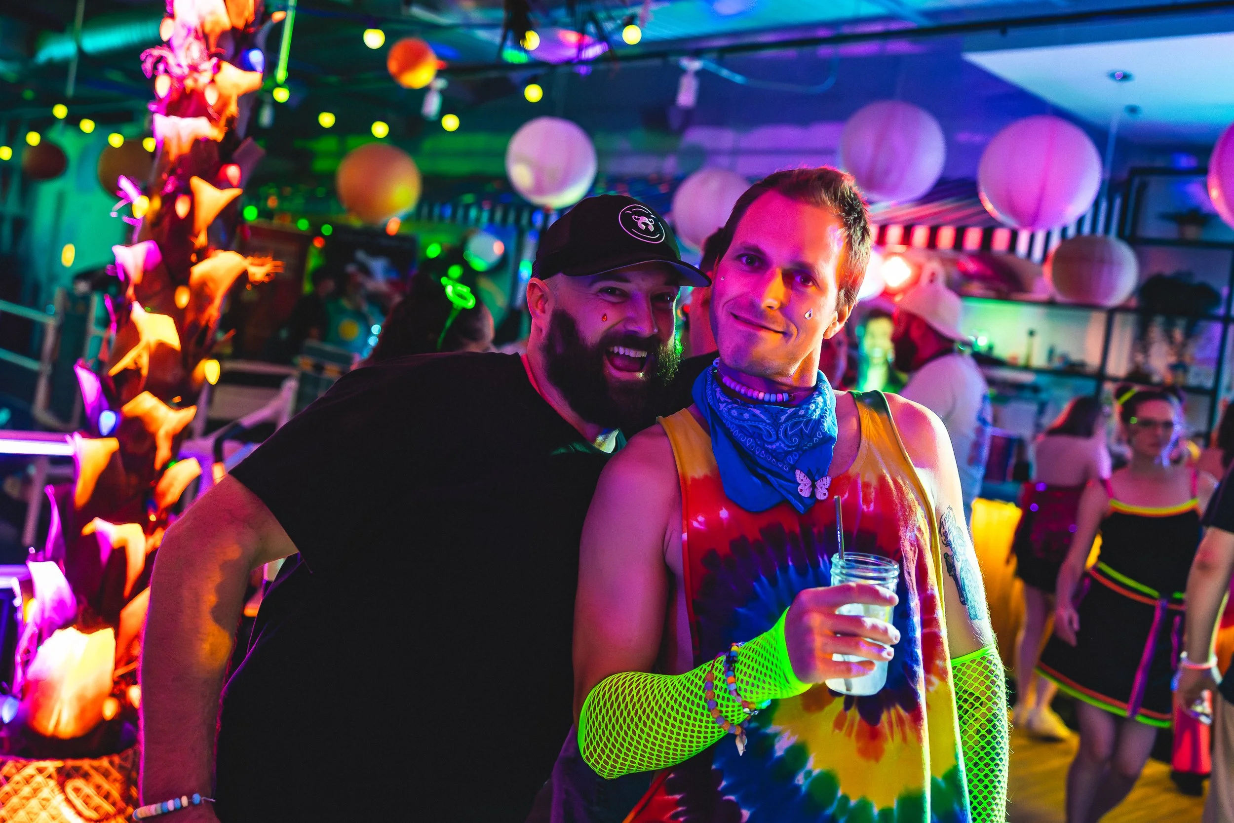 Two men at a colorful, neon-lit party. One man is wearing a black t-shirt and cap, the other is in a tie-dye shirt, neon fishnet arm sleeves, and holding a drink. They are smiling and enjoying the lively atmosphere with hanging paper lanterns and fes