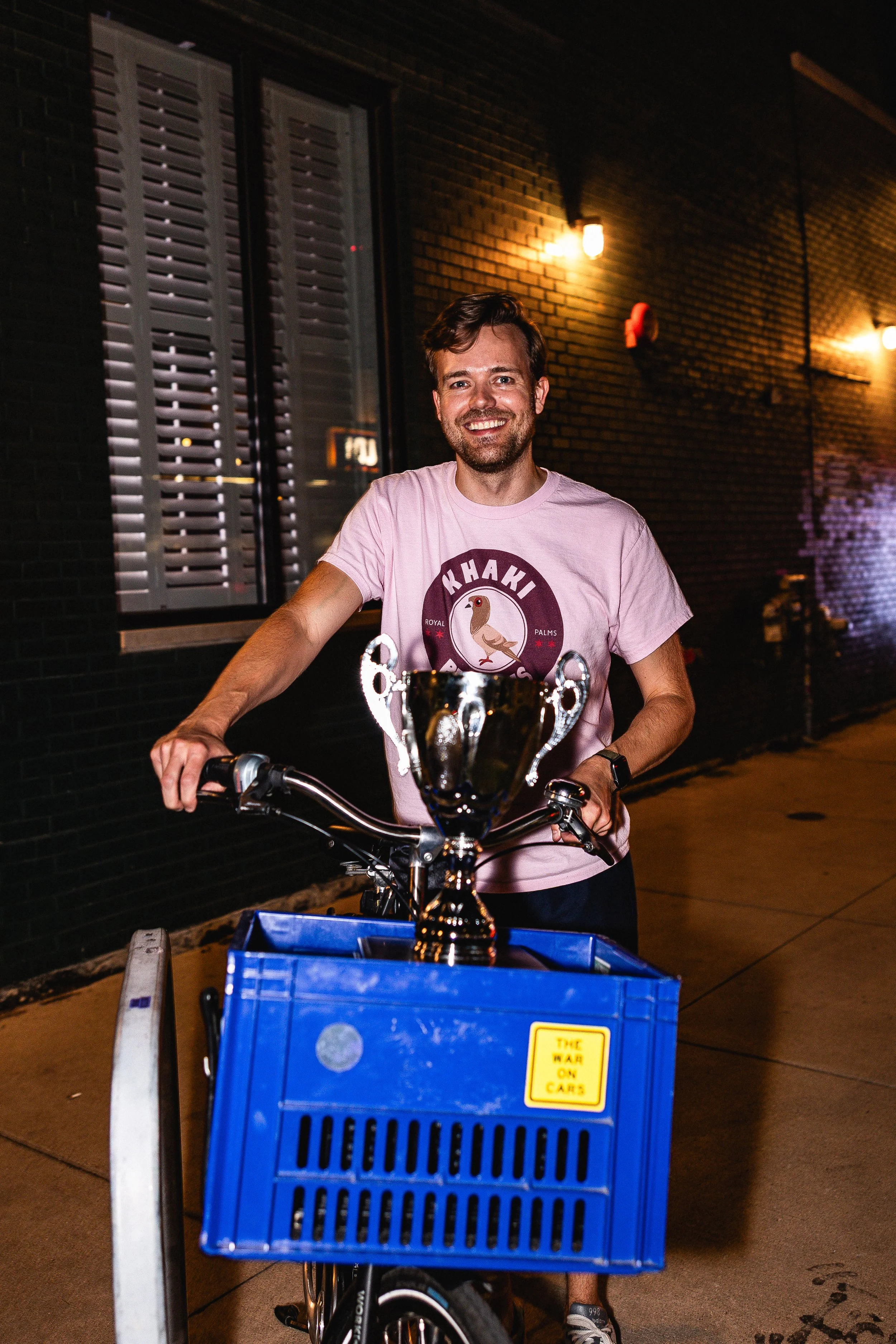 A man smiling and standing on a bicycle with a trophy in a blue crate attached to the front, at night outside a brick building.