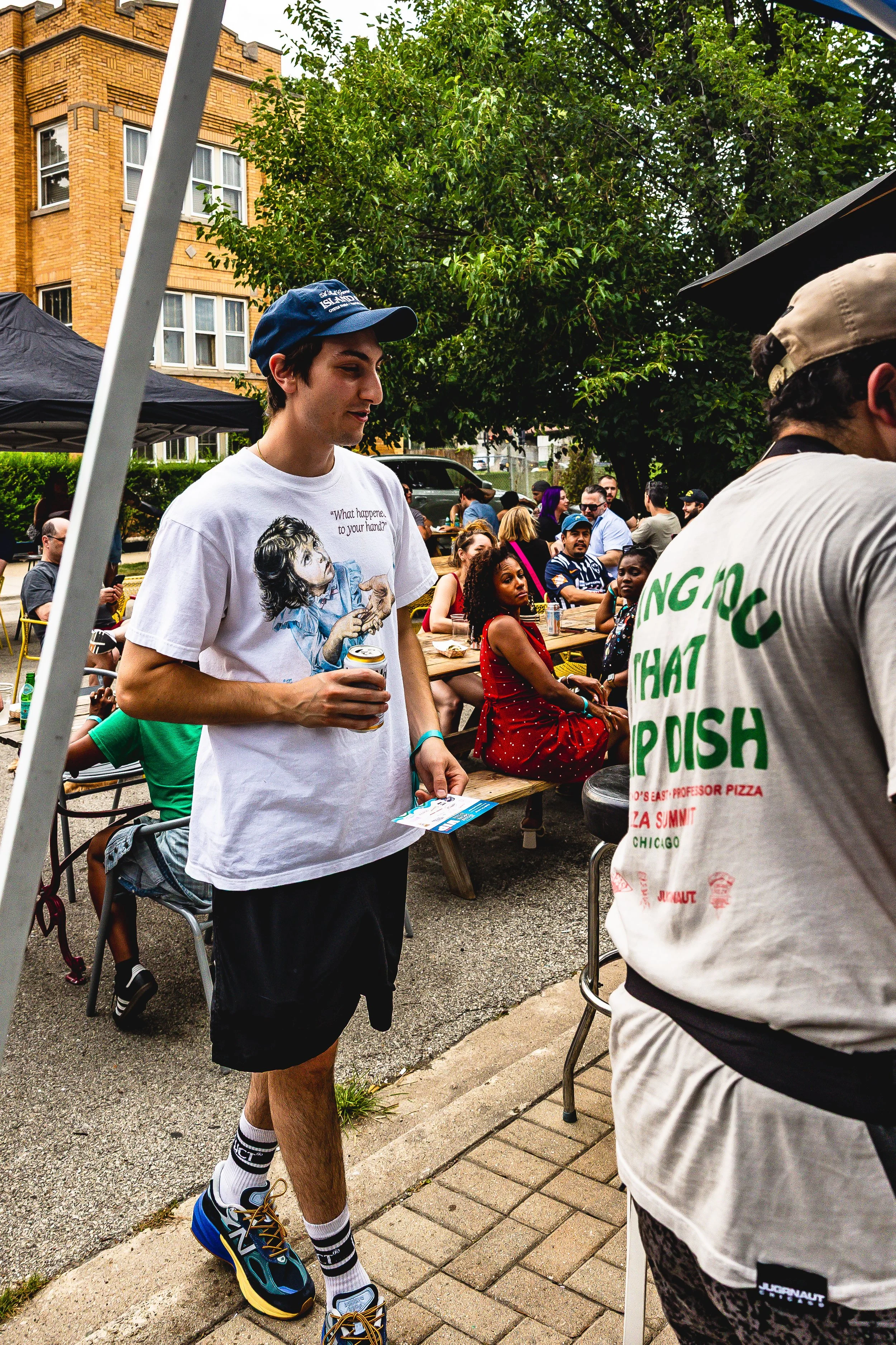 Young man holding a can of beverage at outdoor gathering, wearing a white graphic t-shirt, navy cap, black shorts, and colorful sneakers; other people sitting at picnic tables under tents in a park-like setting with trees and brick buildings.