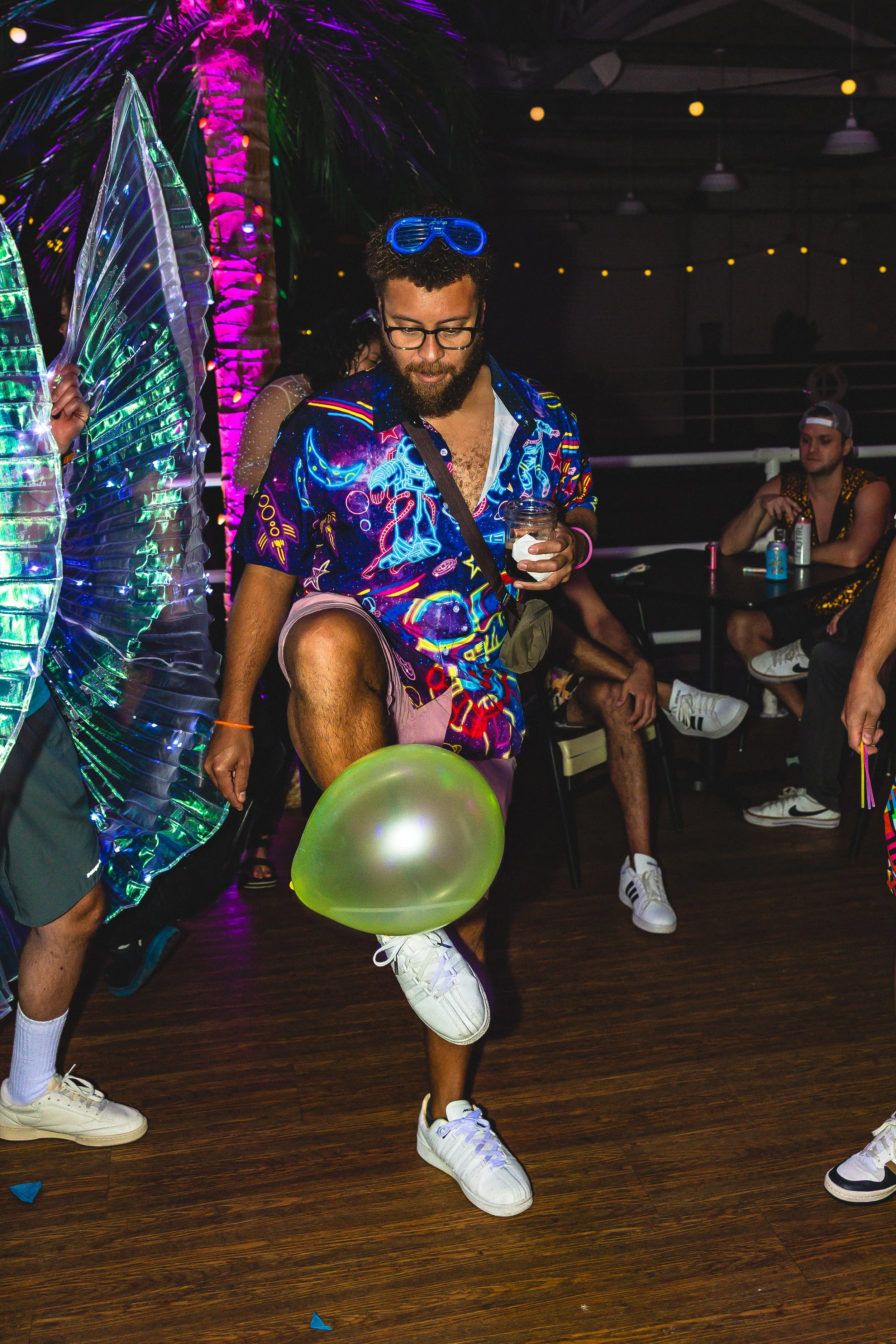 A man at a party wearing a colorful, neon-lit Hawaiian shirt, pink shorts, and white sneakers, holding a drink and balancing a green balloon on one foot. Other guests are seated or standing nearby, and a large illuminated artificial palm tree is in t