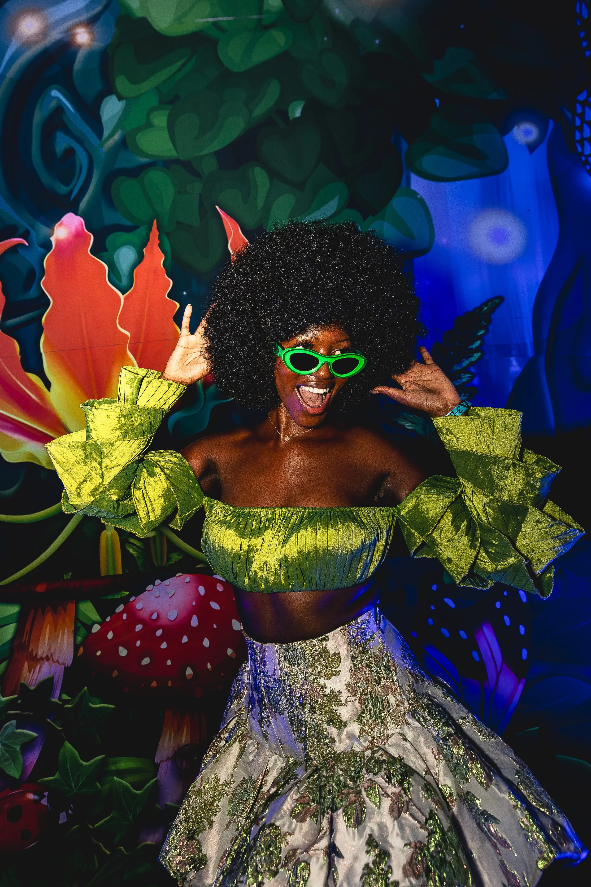 A woman with curly black hair wearing bright green sunglasses, a green off-the-shoulder top with ruffled sleeves, and a patterned skirt, smiling and posing in front of a colorful, psychedelic background with large flowers and abstract designs.