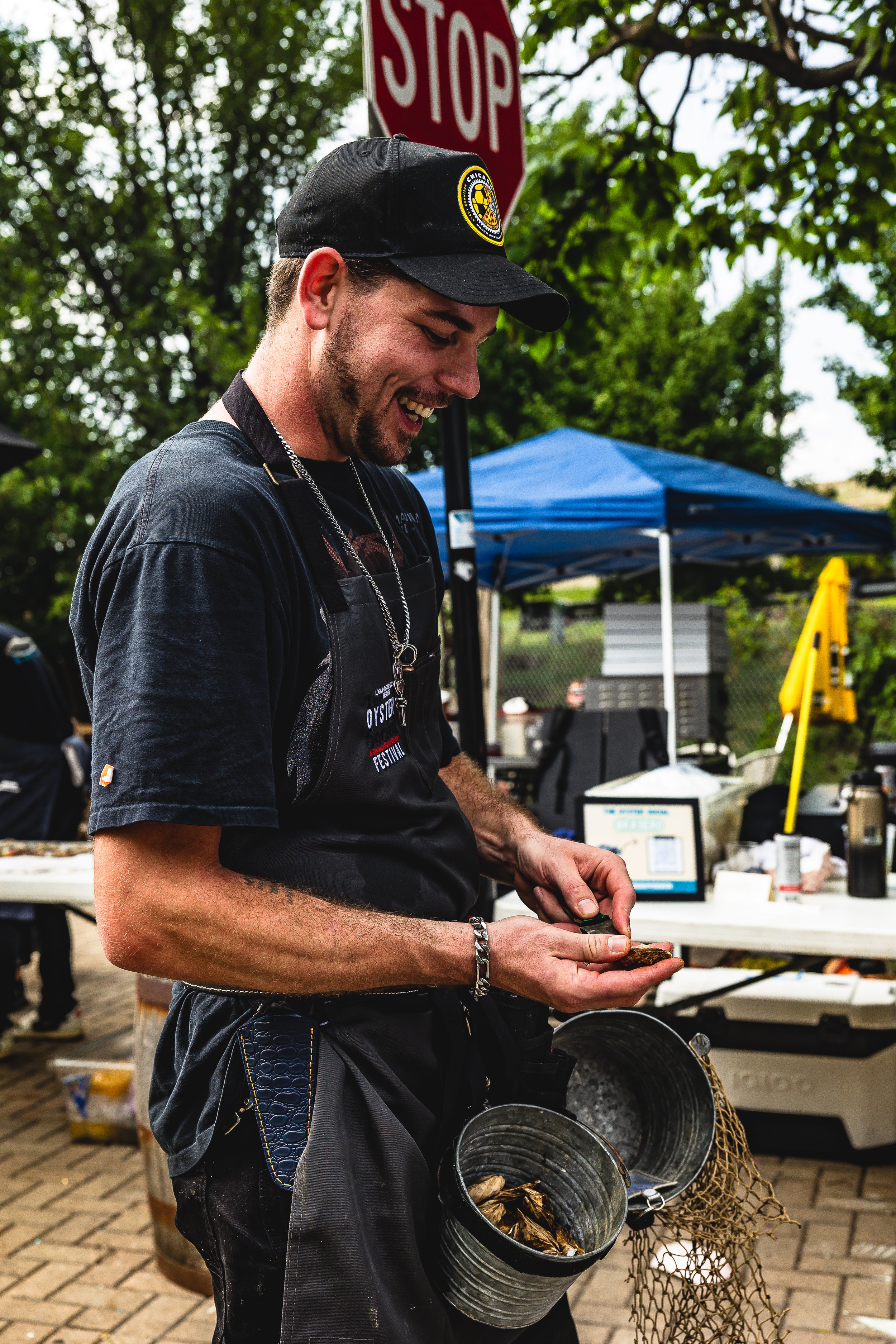 A smiling man with a beard and a black cap examining a shellfish, holding a couple of shellfish in hand, with a bucket of shellfish hanging from his belt, at an outdoor event with tents, a stop sign, and trees in the background.