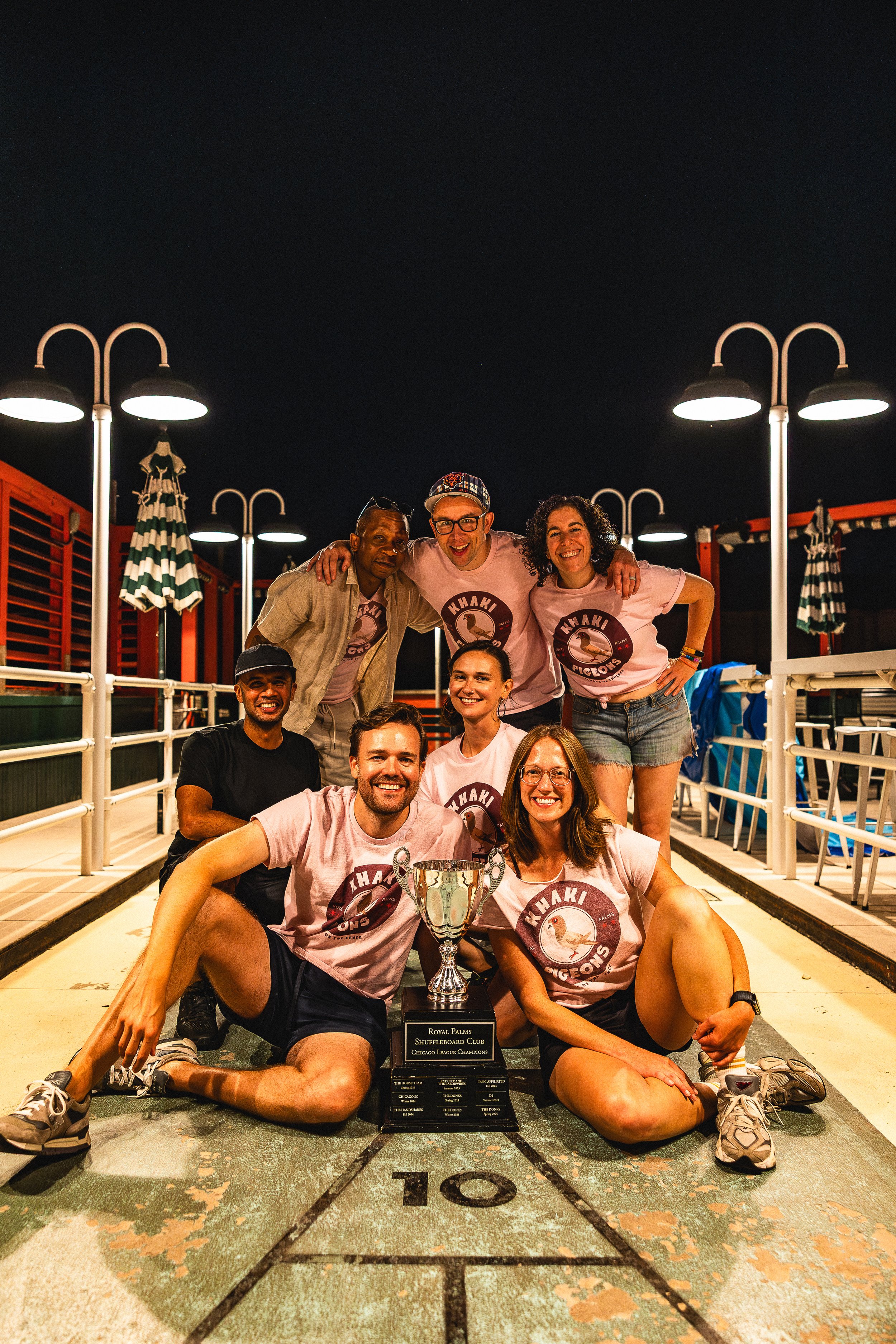 Group of people celebrating a victory on a shuffleboard court at night, posing with a trophy that reads 'Royal Palms Shuffleboard Club, Chicago League Champions.' They are smiling, wearing matching pink shirts with a turtle logo and the text 'Khaki P