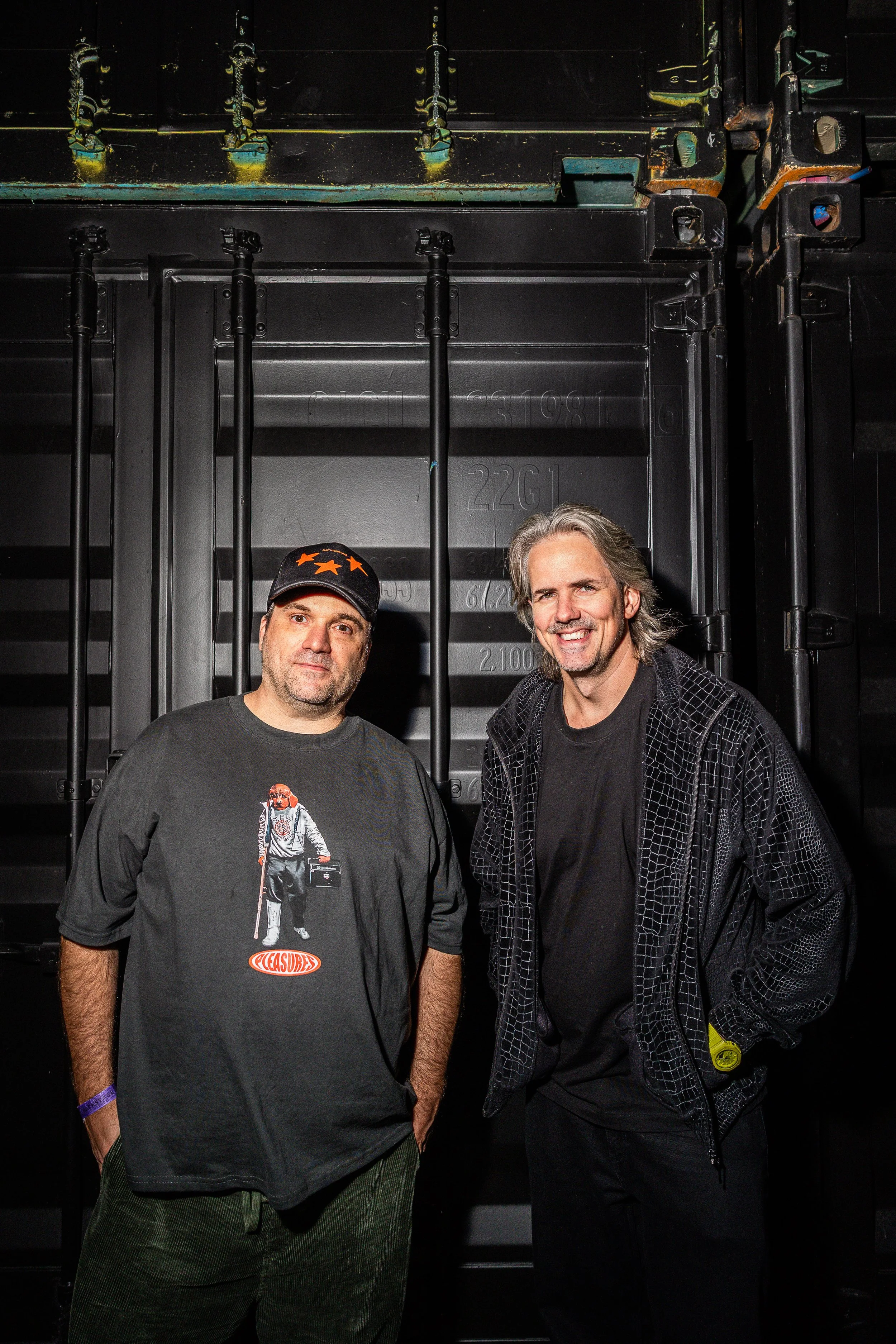 Two men standing in front of a dark, industrial background with equipment. One man is wearing a black T-shirt with a graphic print and a black cap, the other man is smiling and wearing a black jacket.