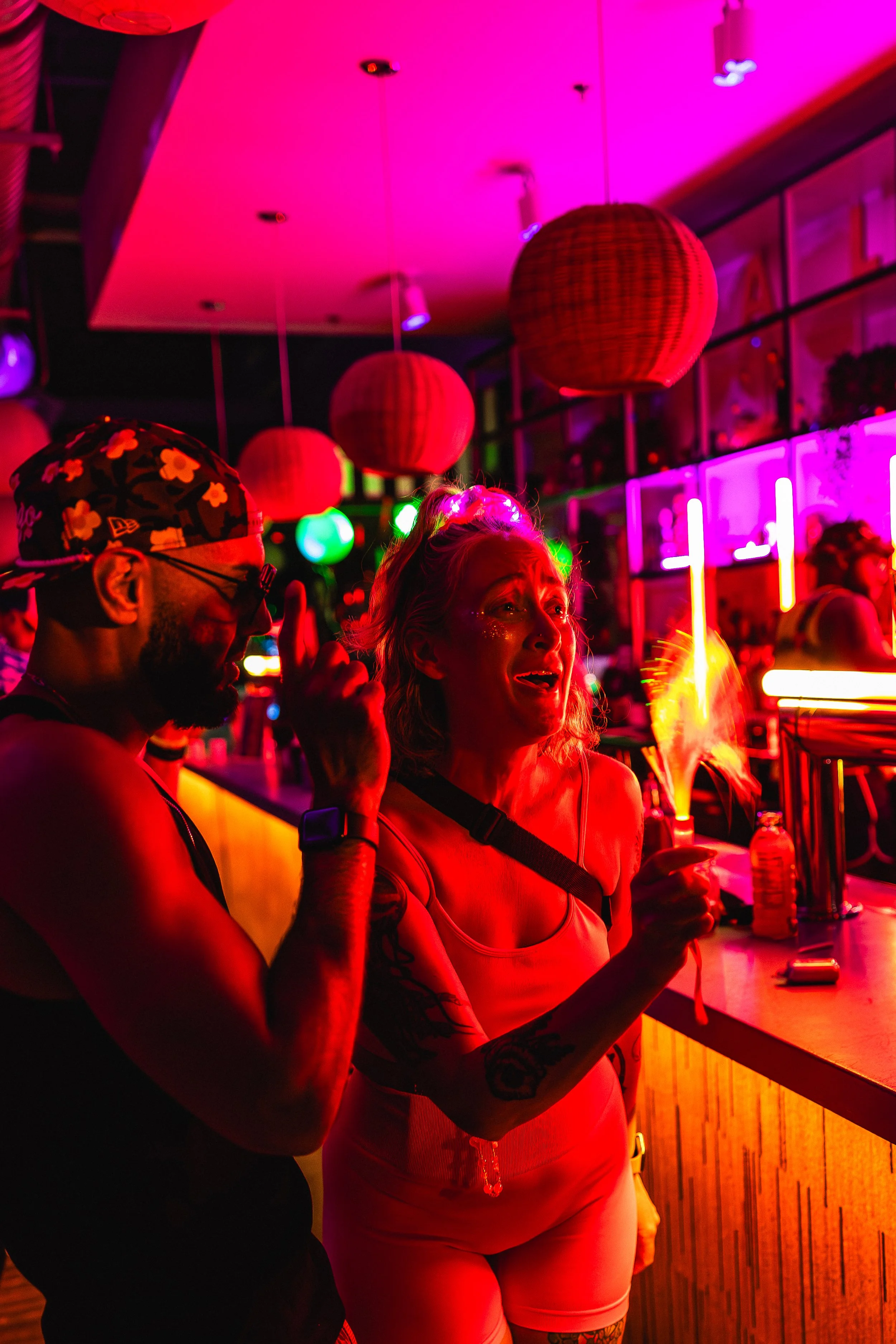 A distressed woman holding a sparkler at a colorful, neon-lit bar or nightclub with a man nearby, illuminated by vibrant pink, green, and purple lights.