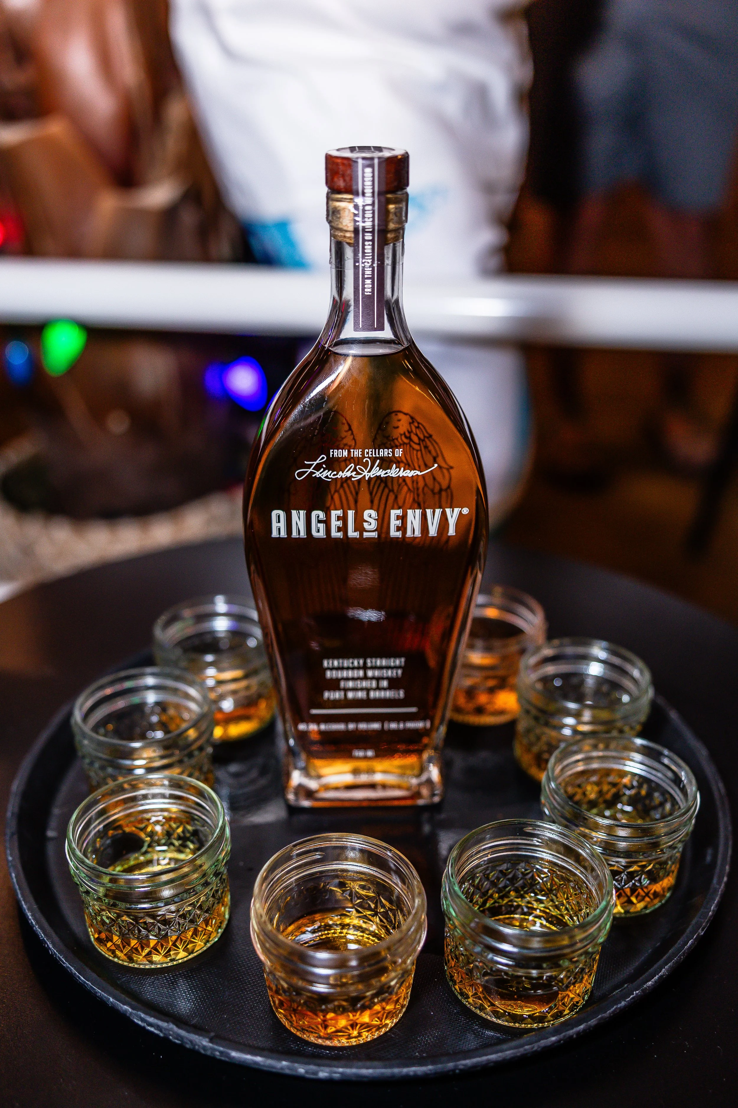 A bottle of Angel's Envy bourbon surrounded by small glasses of bourbon on a round black tray.