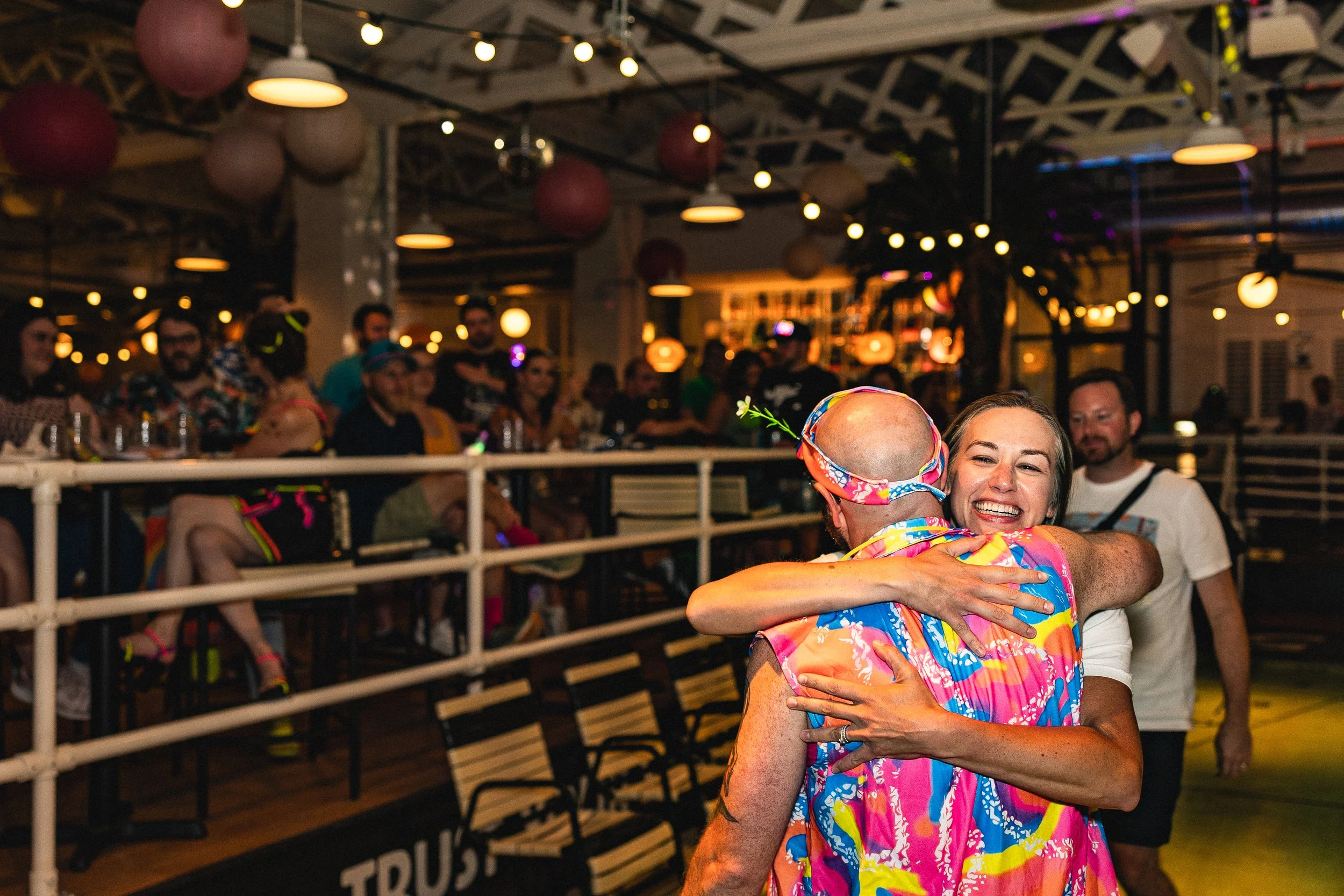 A woman and a man hug joyfully in a lively, decorated social setting with people watching in the background.