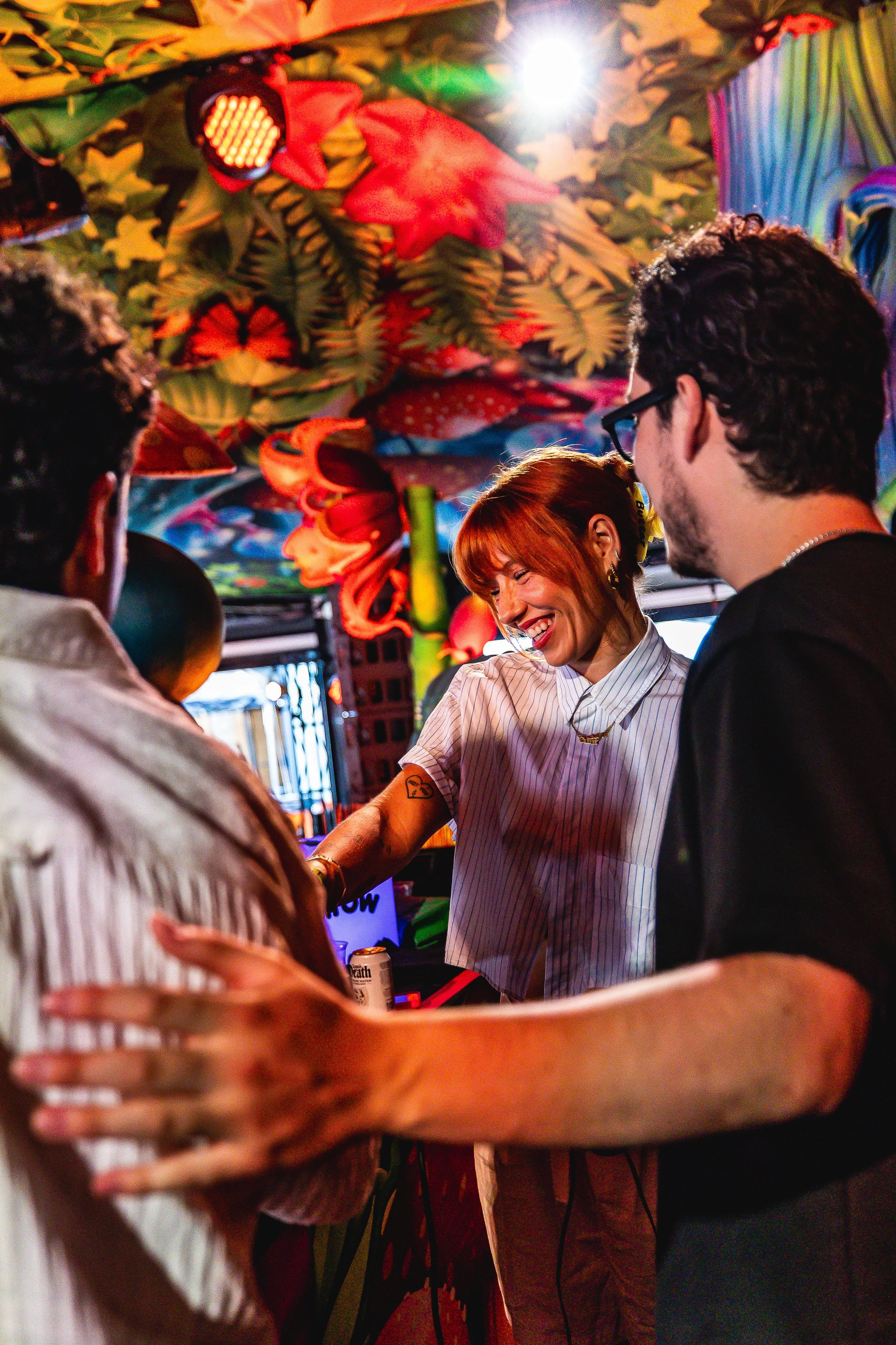 A woman with red hair and tattoos laughing and dancing at a colorful, tropical-themed party with two men.
