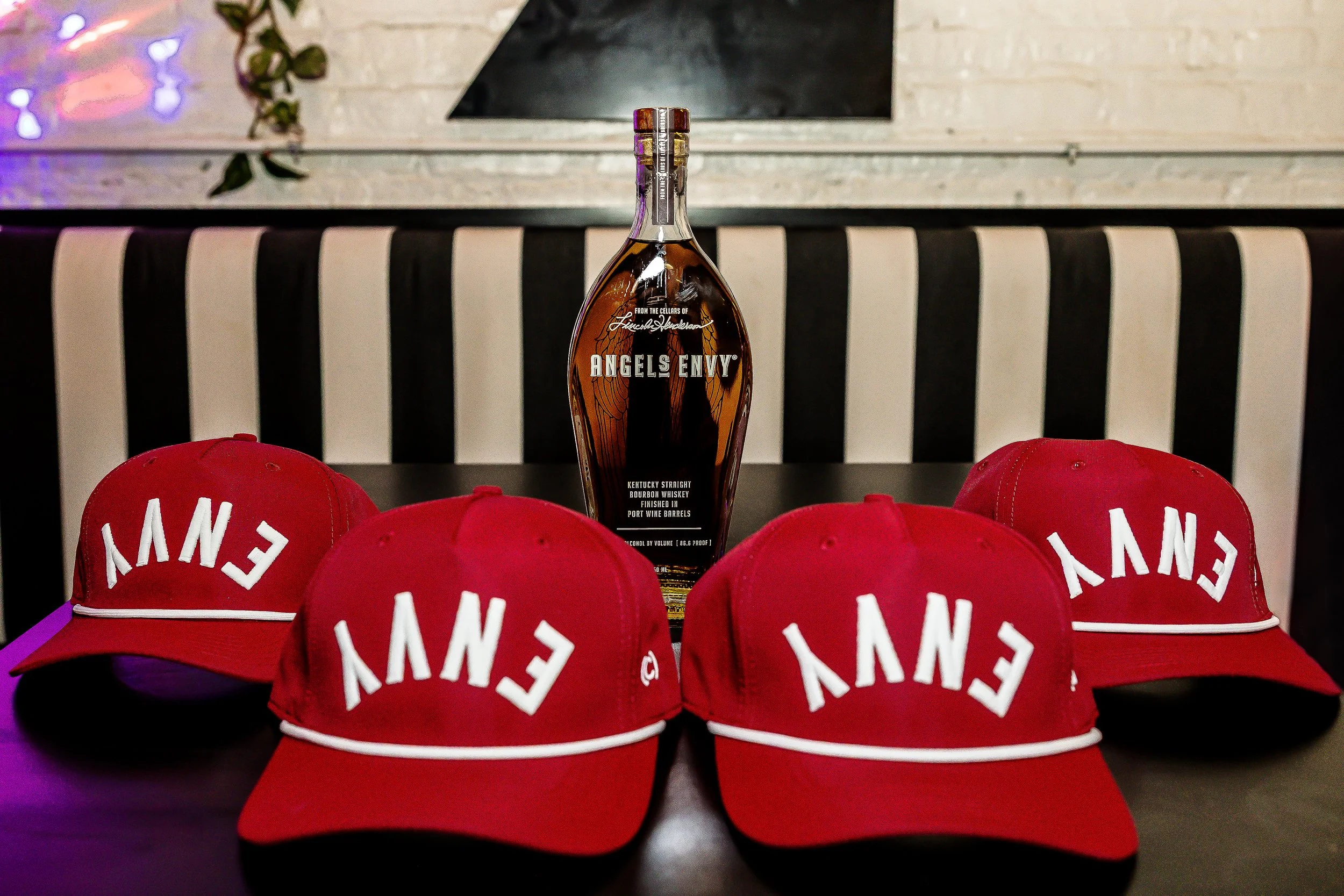 Four red baseball caps with white embroidered letters spelling 'ENVY' are arranged on a black surface. In the background, there is a bottle of Angels Envy bourbon whiskey on a table, with a black and white striped wall behind it.