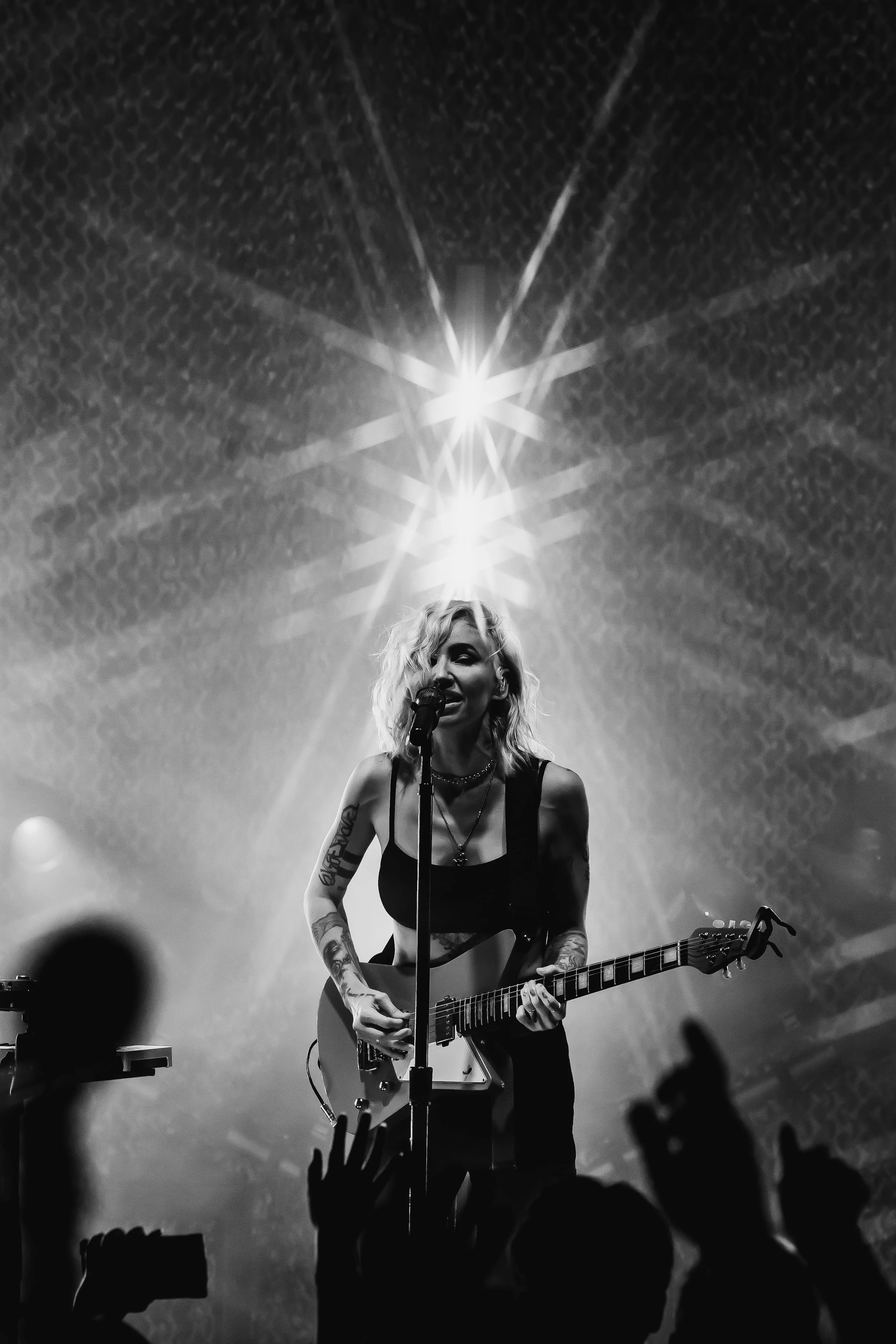 A female musician performing on stage with a guitar, singing into a microphone, surrounded by stage lights and an audience silhouette.