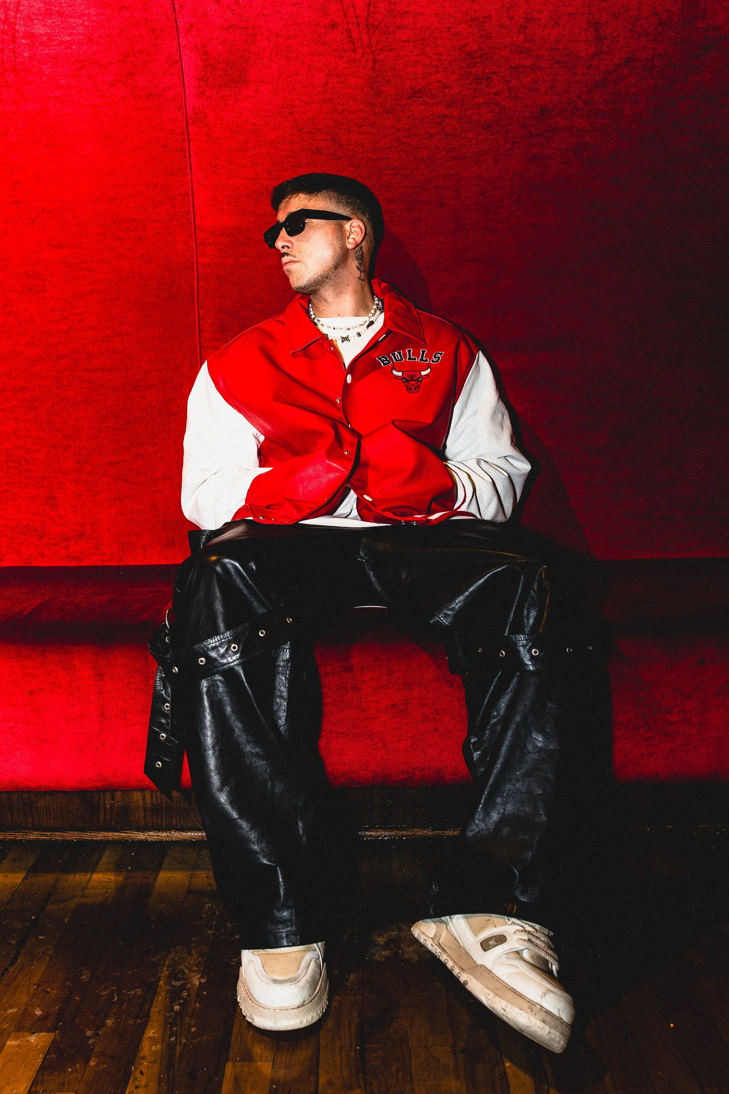 Matroda man sitting on a red bench against a red background, wearing sunglasses, a red and white varsity jacket with 'BULLS' and a bull logo, baggy black leather pants with metal eyelets, and white sneakers.
