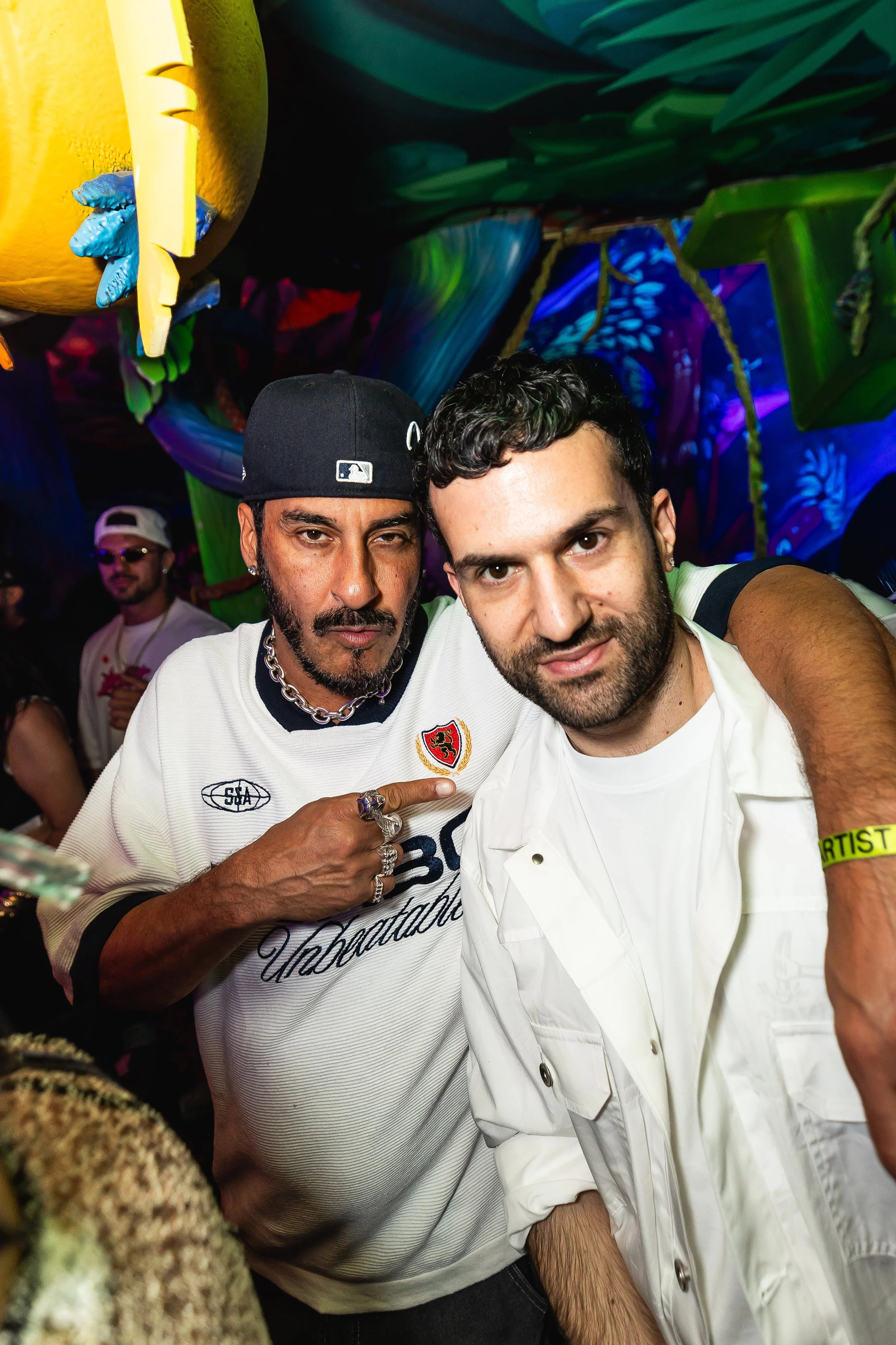 Two men taking a selfie at a crowded party or nightclub with colorful, psychedelic lighting on a vibrant ceiling