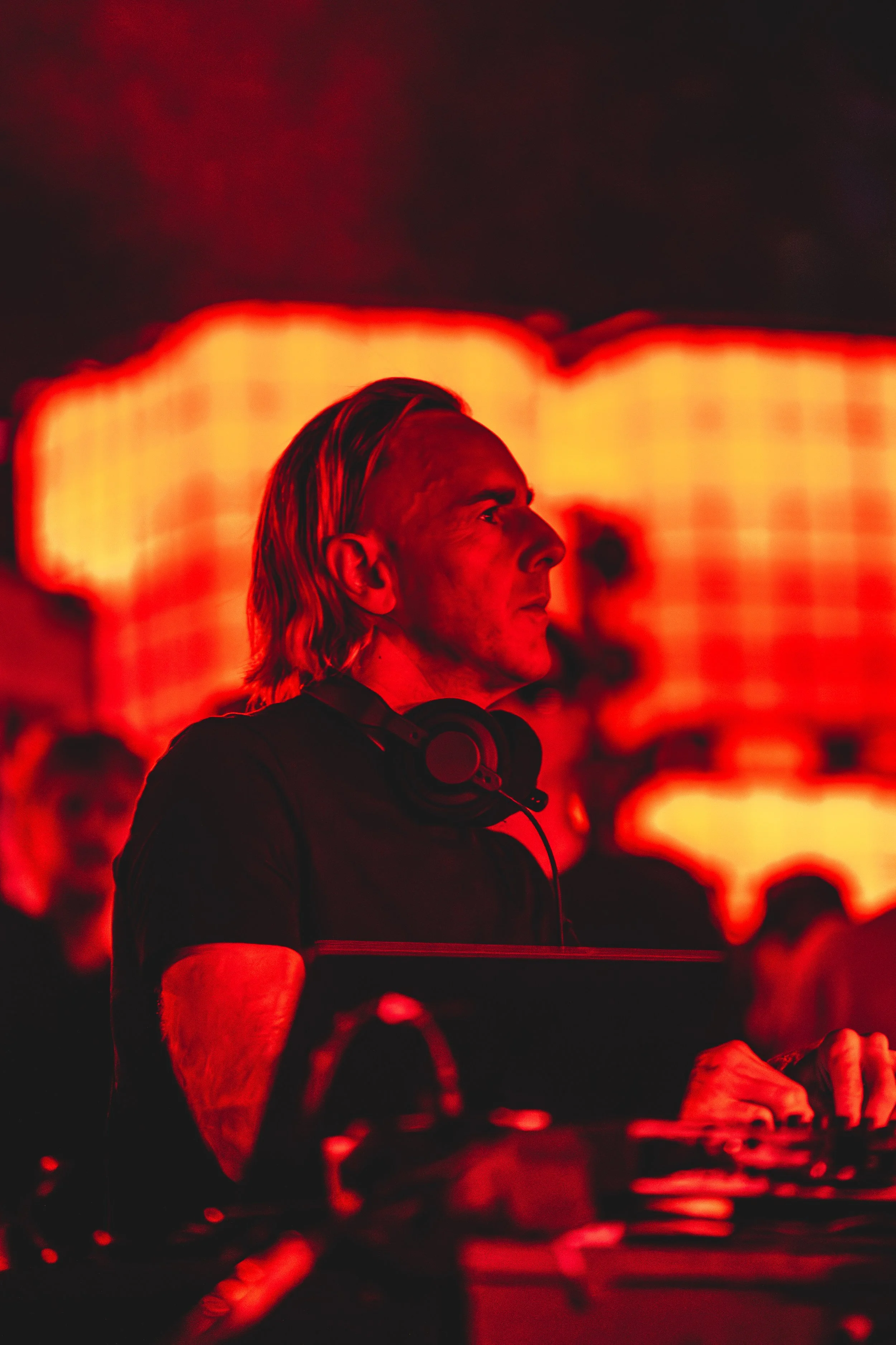 A DJ with shoulder-length blonde hair and headphones around his neck, working at his equipment with a fiery red and orange LED background.