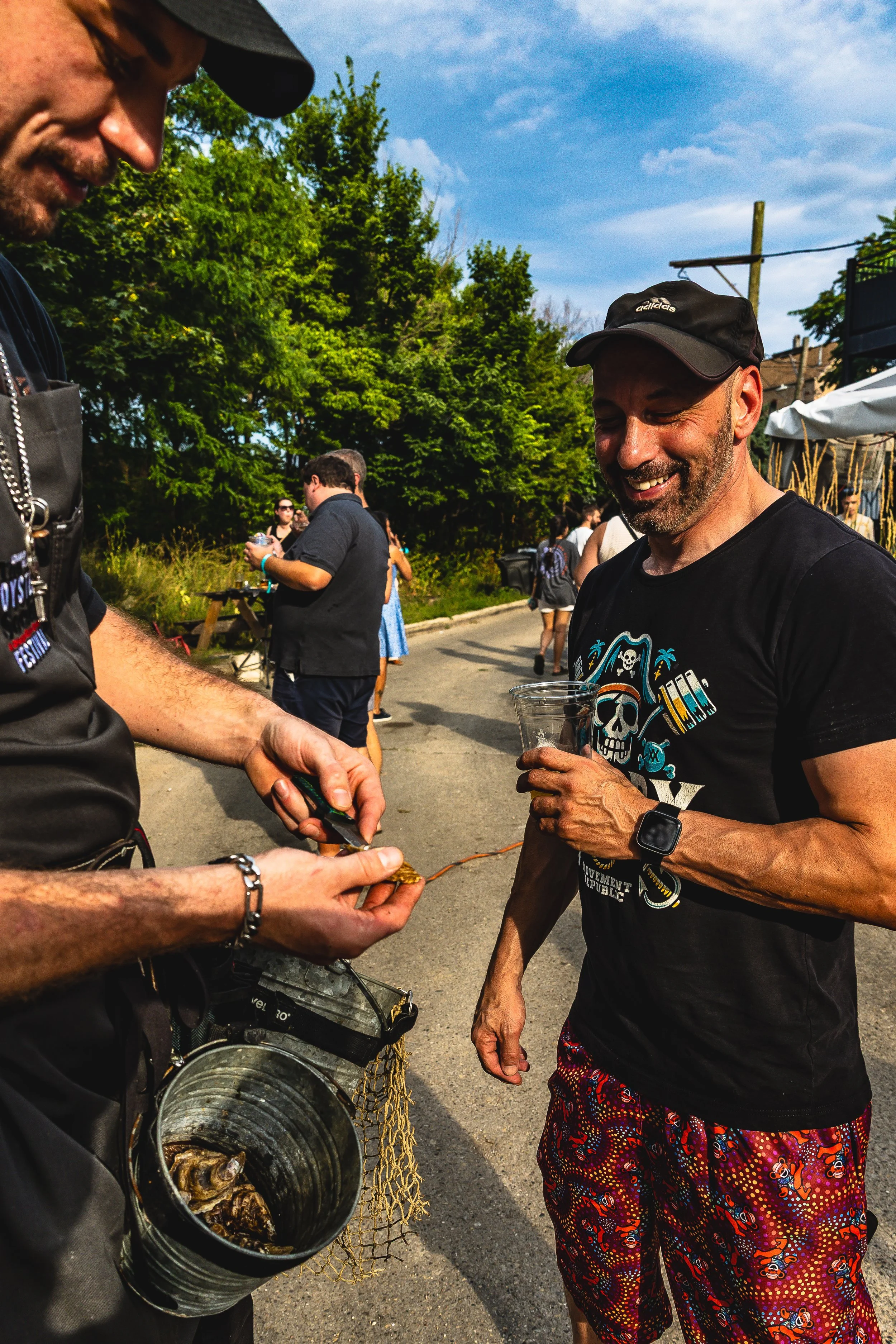 Two men at an outdoor event, one holding a plastic cup, the other using pliers on a small object, with several people and trees in the background.