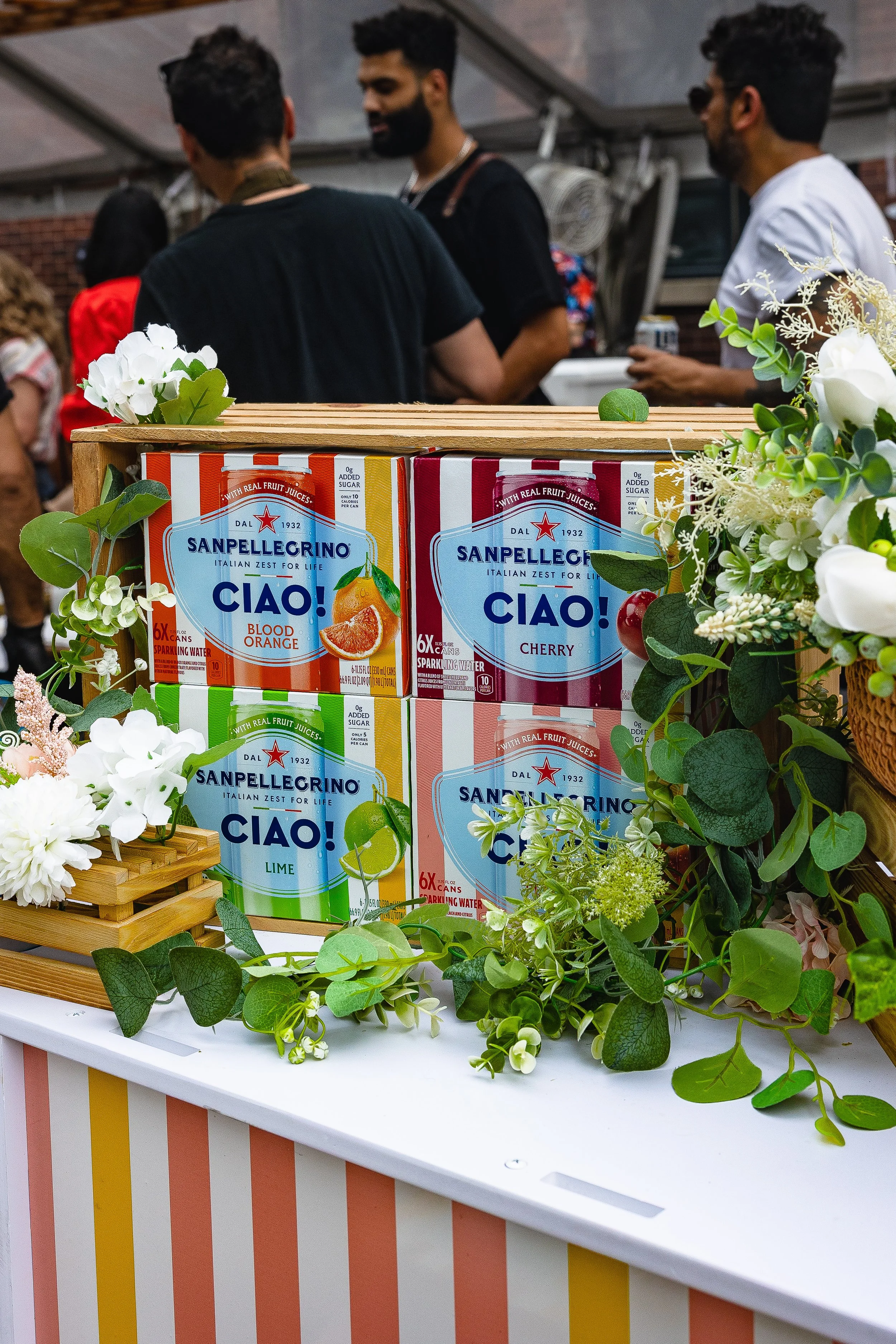 Display of Sanpellegrino Ciao! Italian fruit juices with flavors of blood orange, cherry, and lime, decorated with flowers and greenery, at an outdoor event with people in the background.