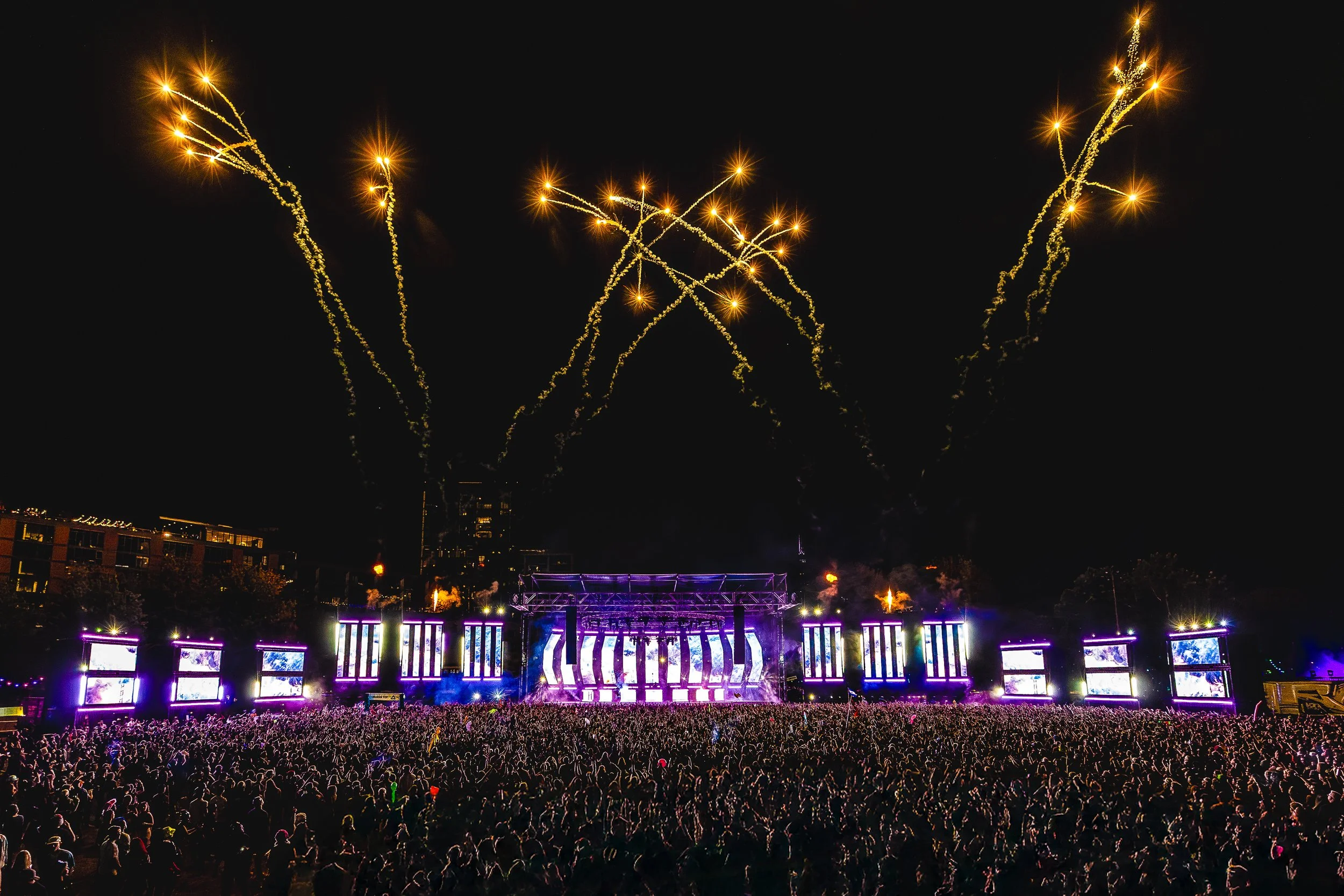 A large crowd at a nighttime outdoor music festival with fireworks in the sky and a brightly lit stage with multiple screens and colorful lights.