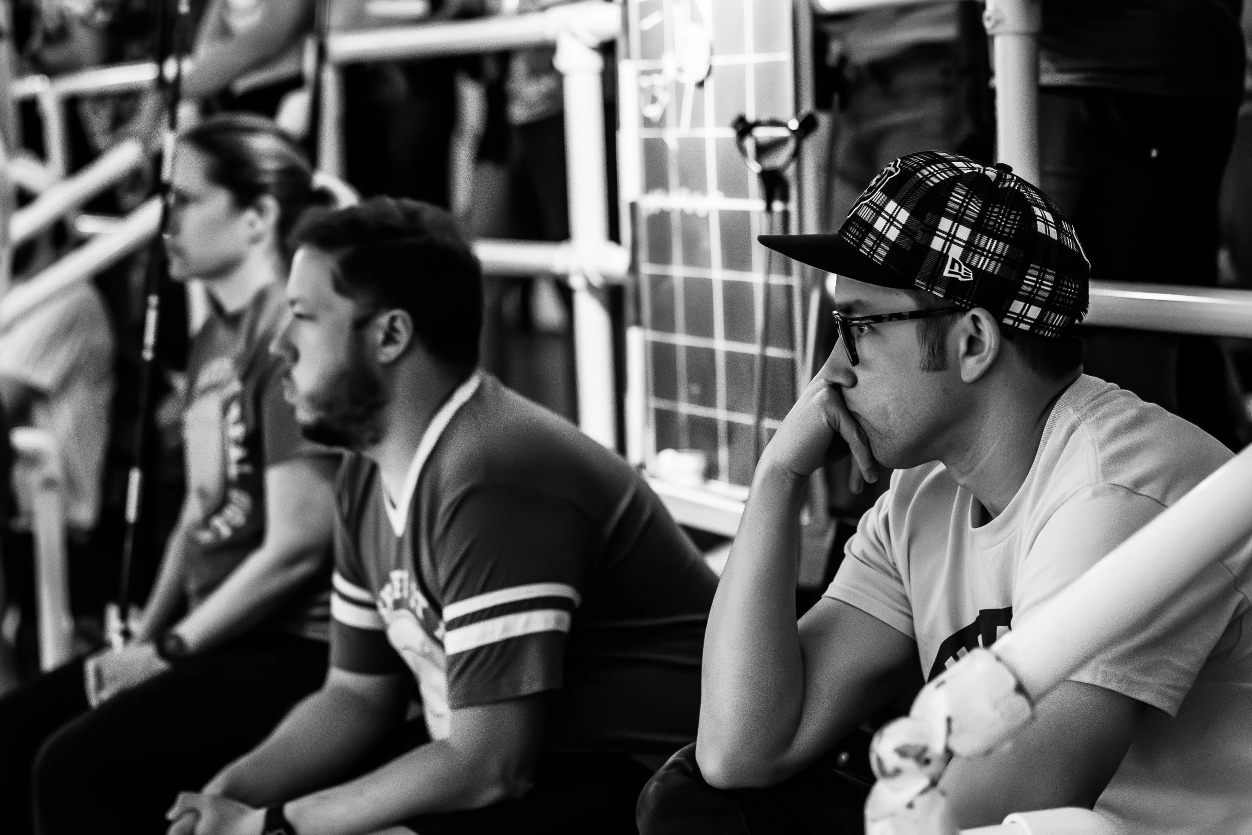 Four people sitting in a row, appearing to be at an event or game, with two women and two men, one wearing a plaid cap and glasses, all looking attentively forward in a black and white photo.