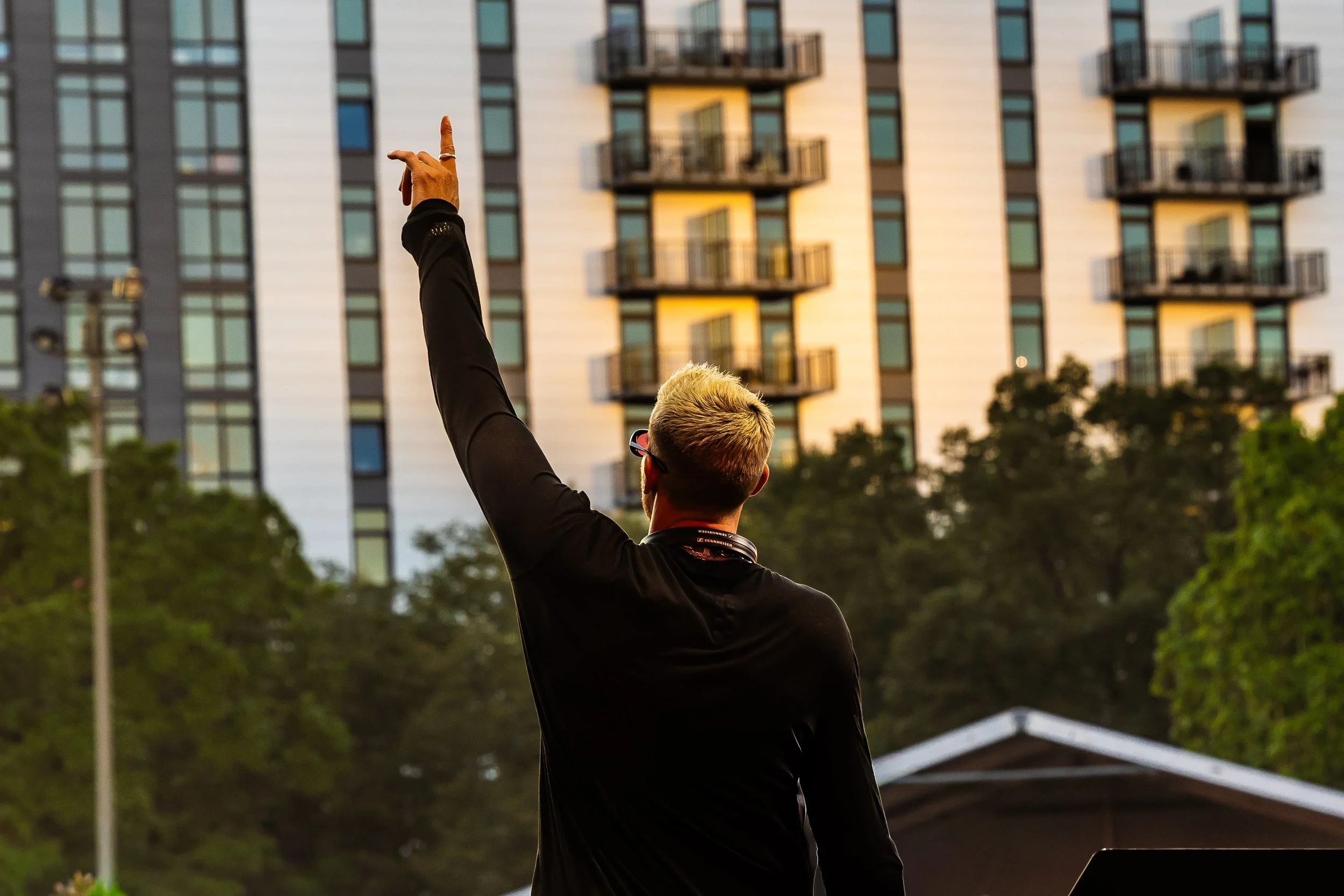 A person with blonde hair and glasses, wearing a black shirt, standing outdoors in front of a tall building with balconies, raising one arm with a finger pointing upward.