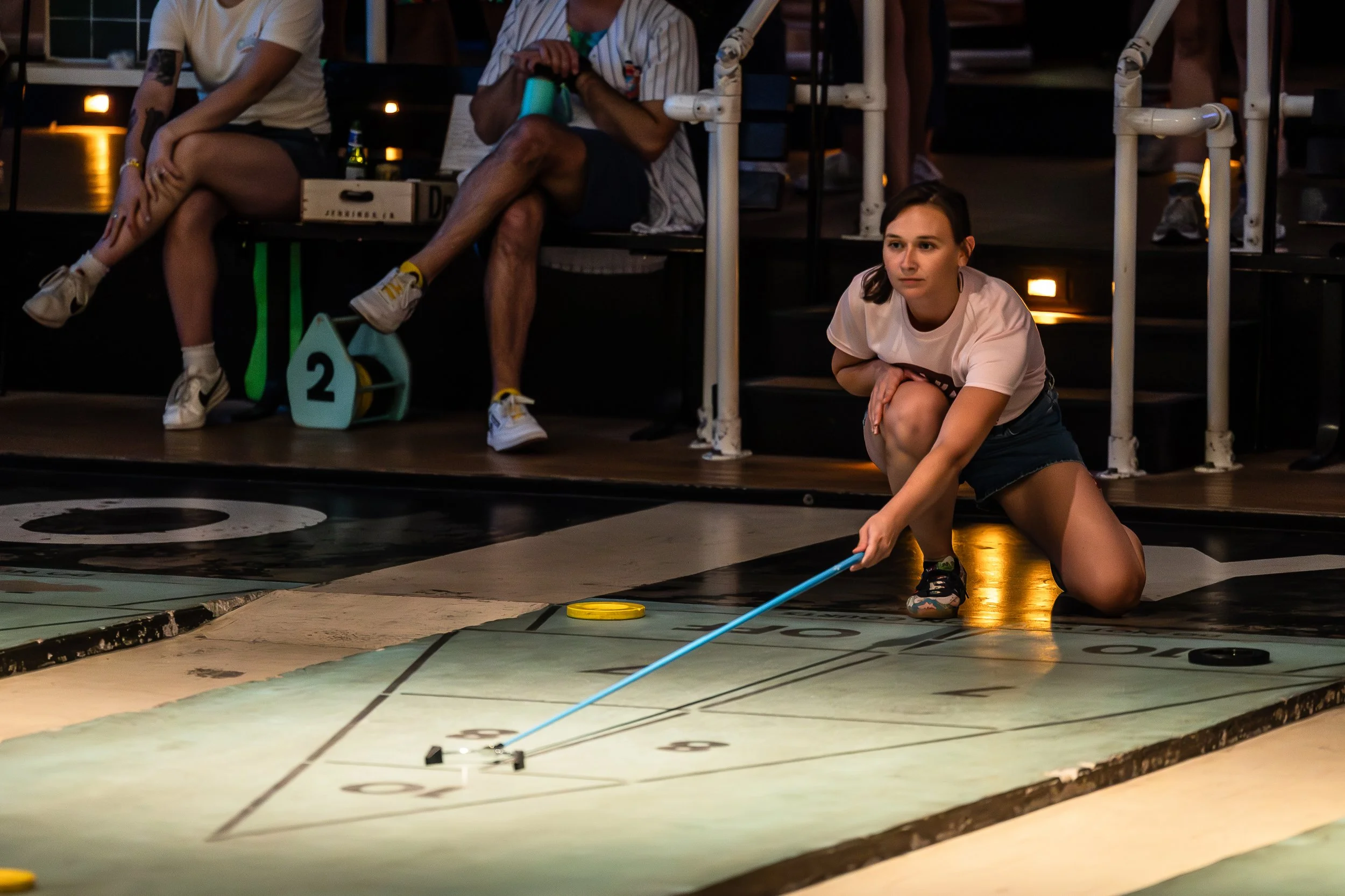 A young woman is crouching on the floor, playing shuffleboard with a cue and puck, indoors at a recreational facility.