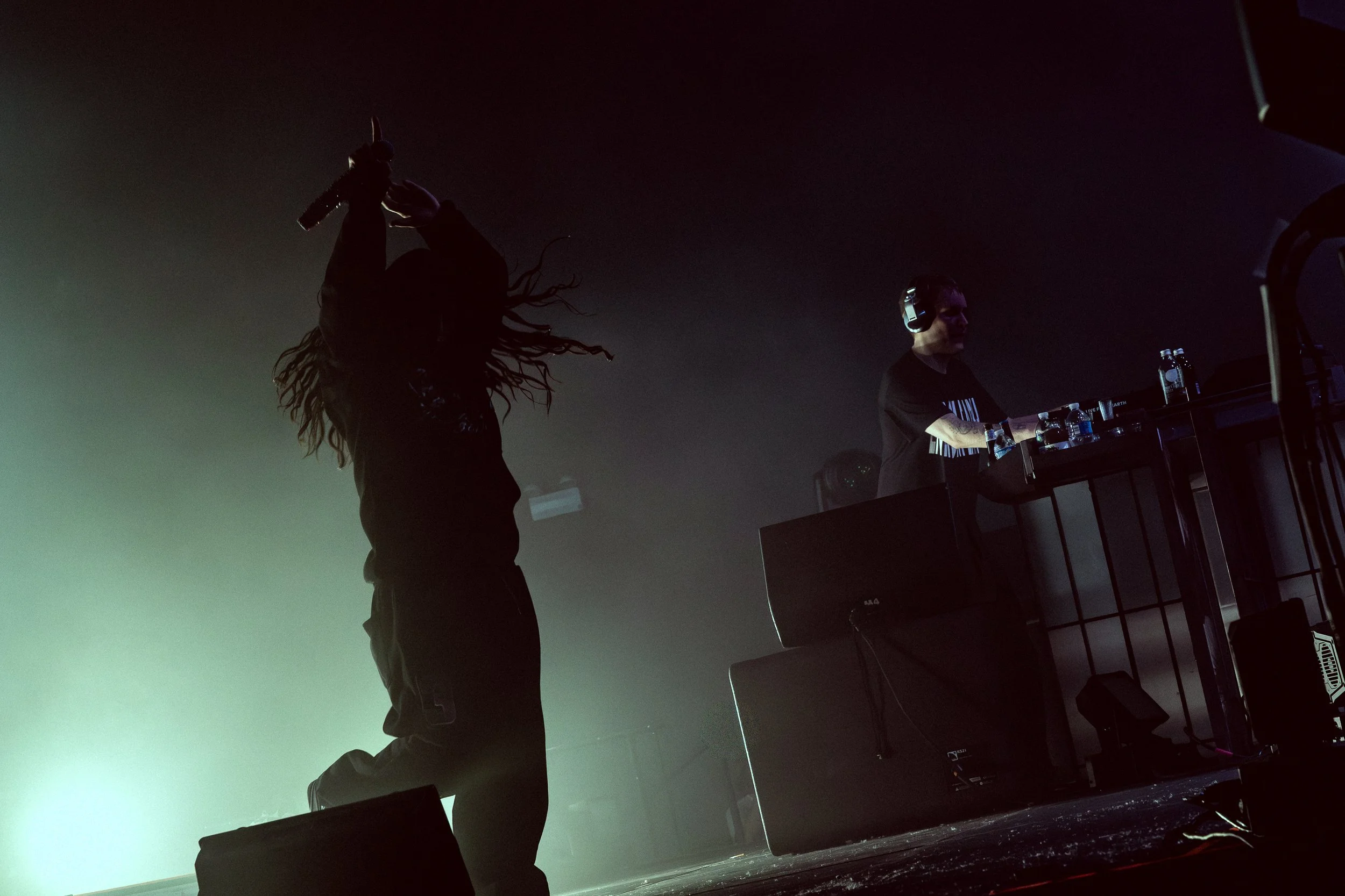 A singer with dreadlocks performing on stage with a microphone, and a DJ wearing headphones and using a mixer, in a dimly lit concert setting.