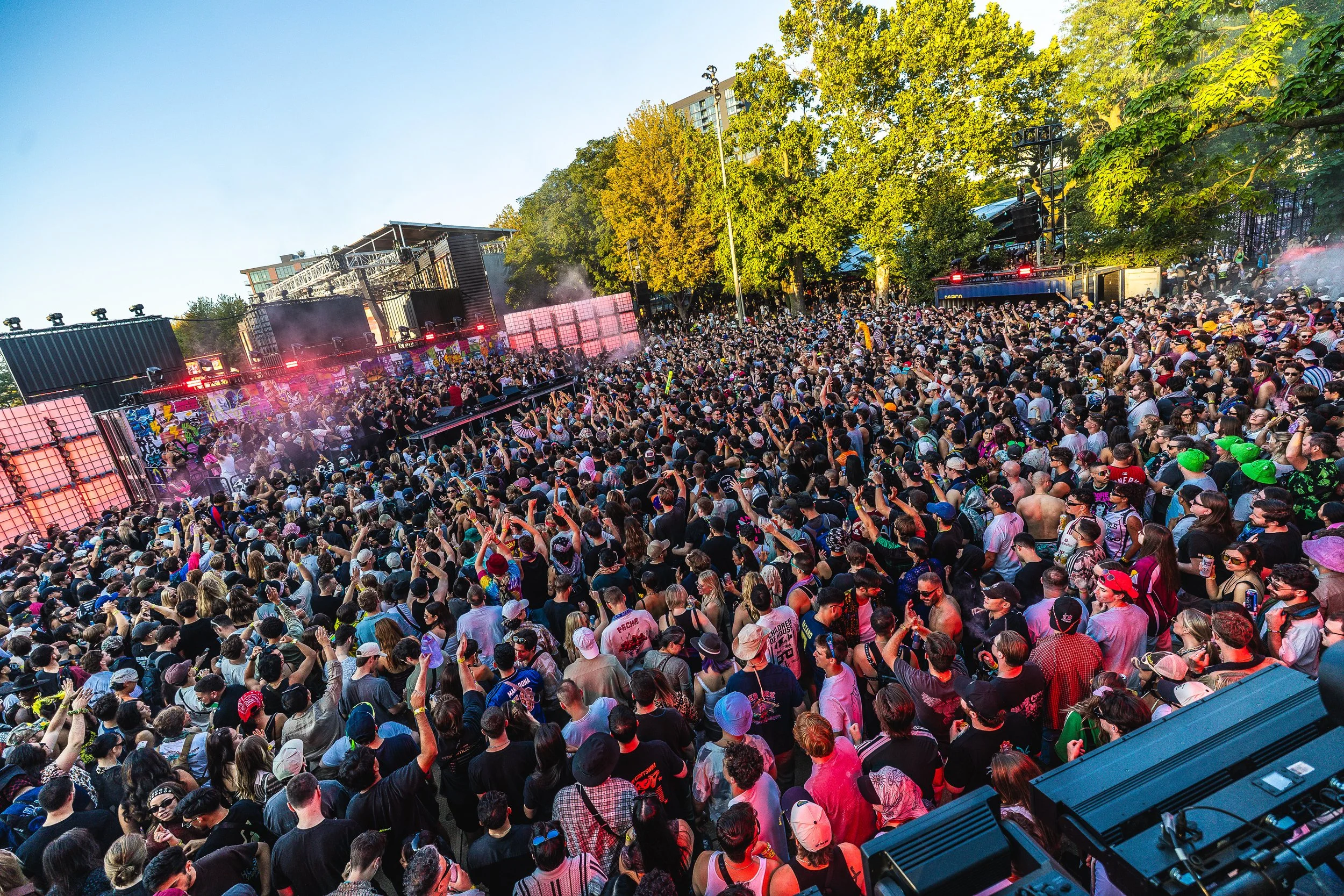 A large crowd of people at an outdoor music concert, with a stage and vibrant lighting, surrounded by trees and clear sky.