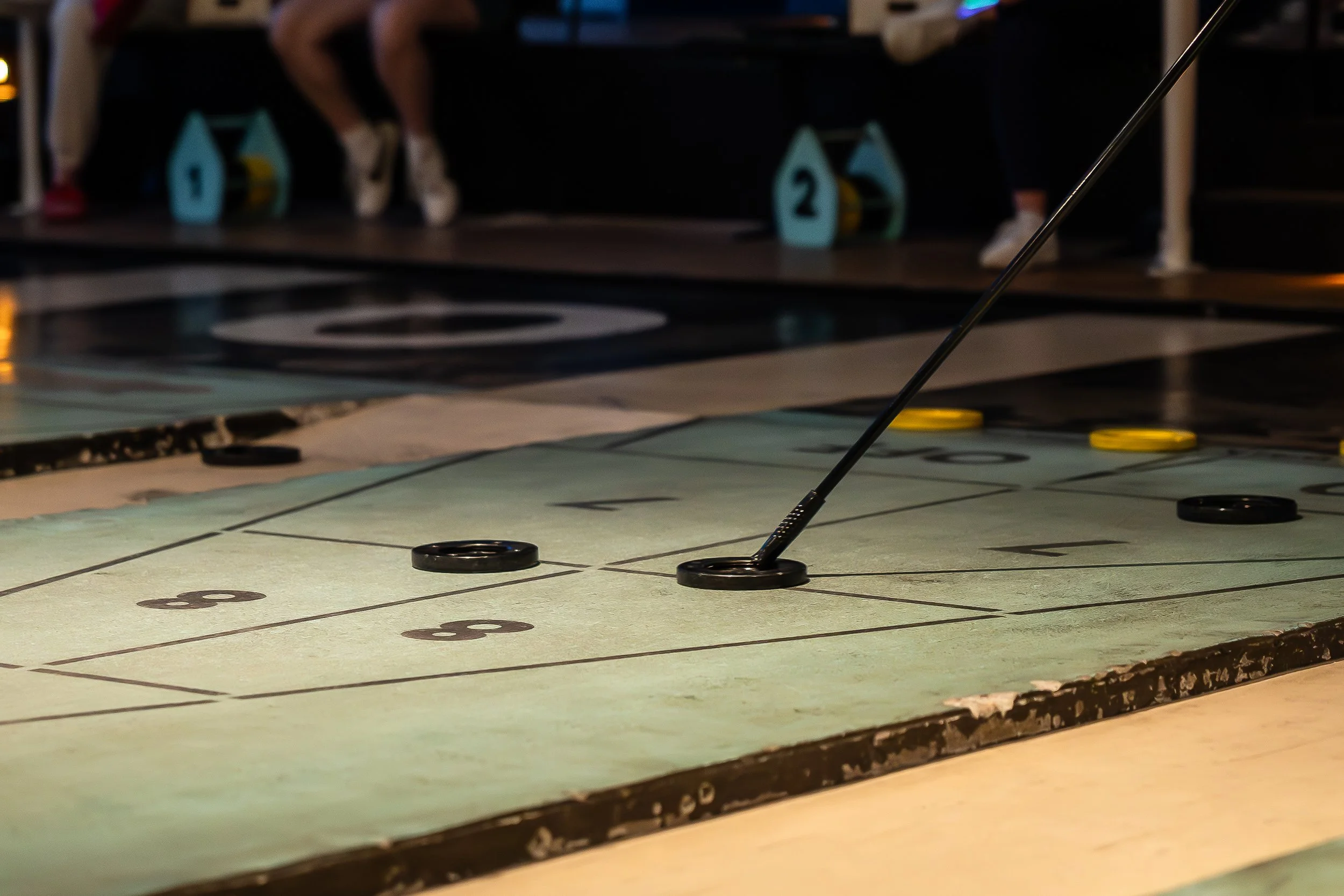 A floor game of shuffleboard with pucks and a cue stick, with players in the background.