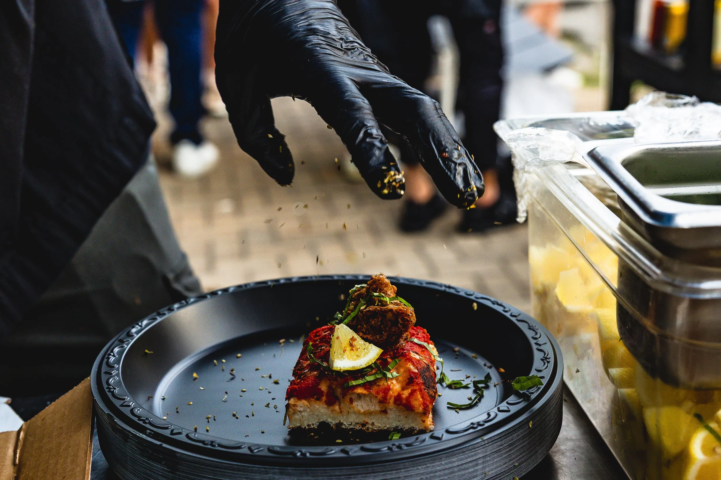 Person wearing black gloves sprinkling herbs or spices on a slice of pizza with toppings of cheese, tomato sauce, herbs, and a lemon wedge, on a black plate.