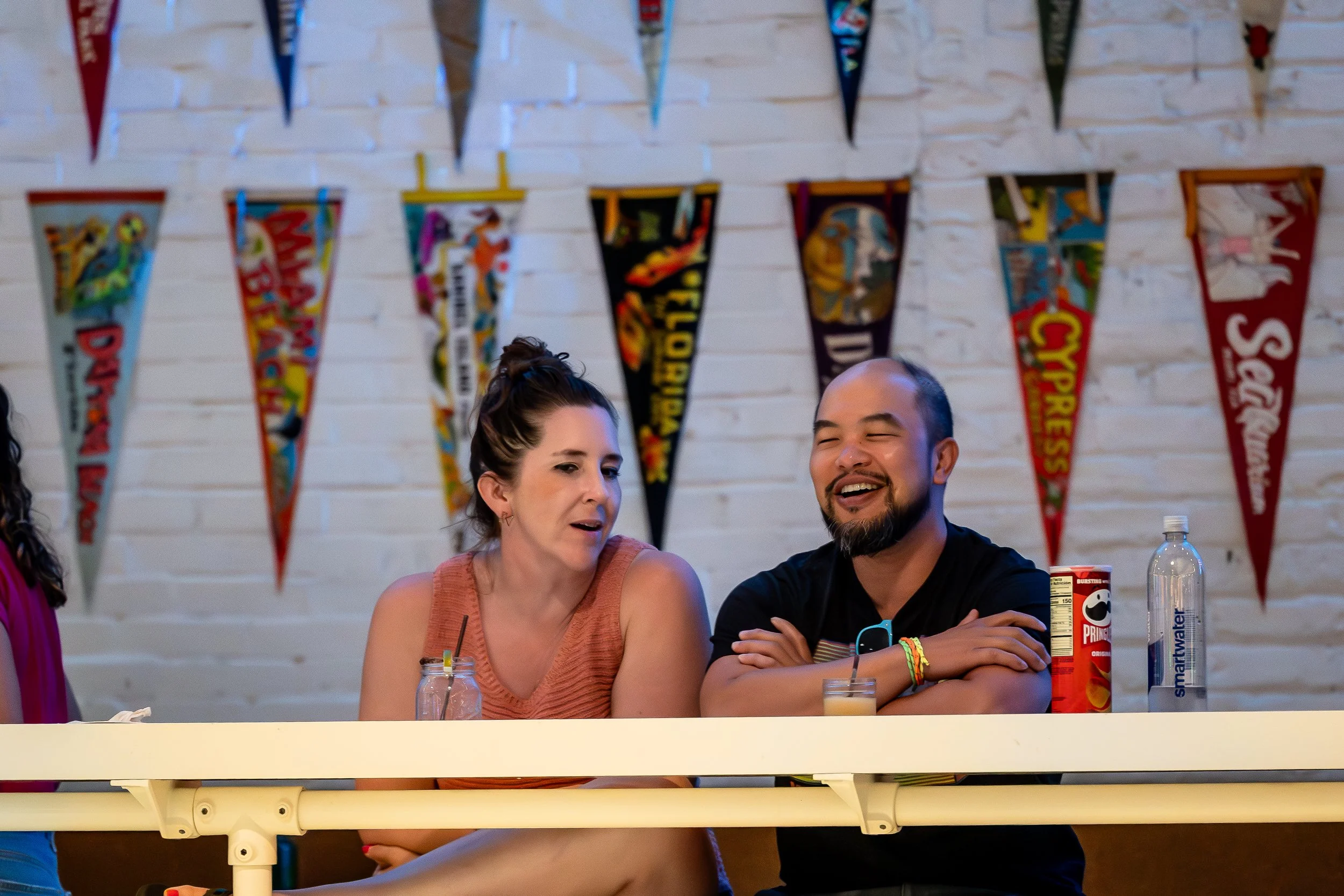 Two people sitting at a table, smiling and talking, with colorful pennant banners on a white brick wall behind them. One woman has dark hair in a bun wearing an orange top; one man has a beard and buzz cut, wearing a black t-shirt.