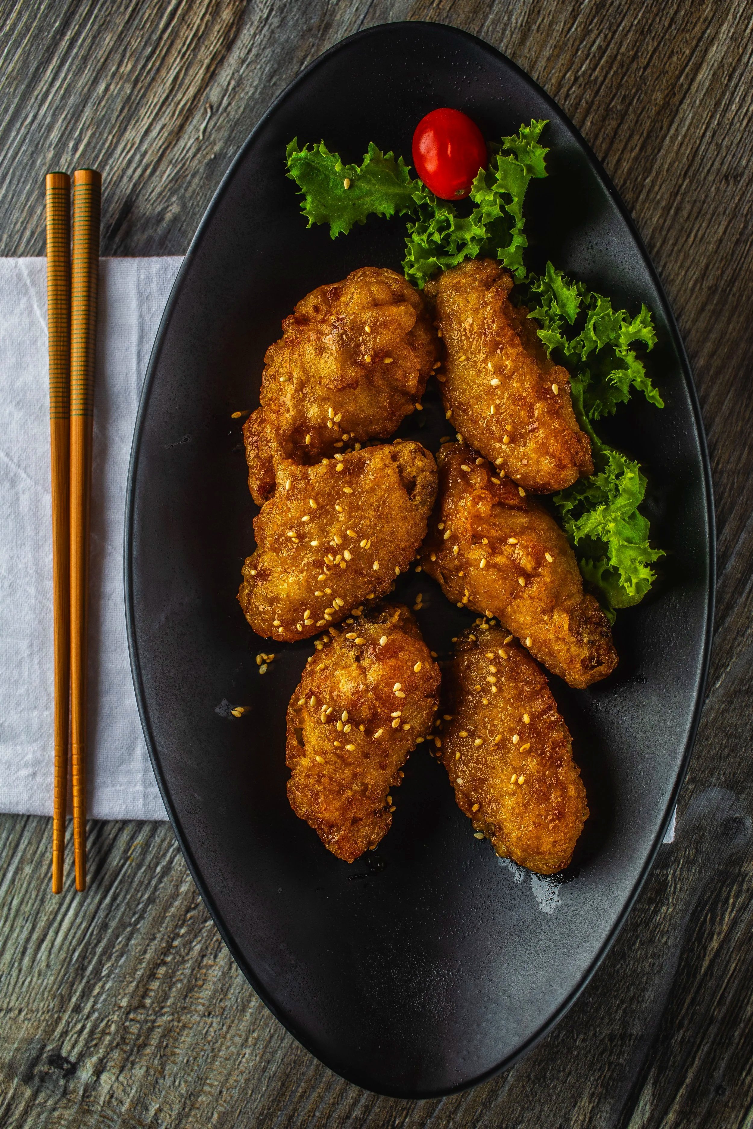 Fried chicken wings sprinkled with sesame seeds on a black oval plate, garnished with leafy greens and a cherry tomato, with chopsticks resting on a napkin beside the plate.