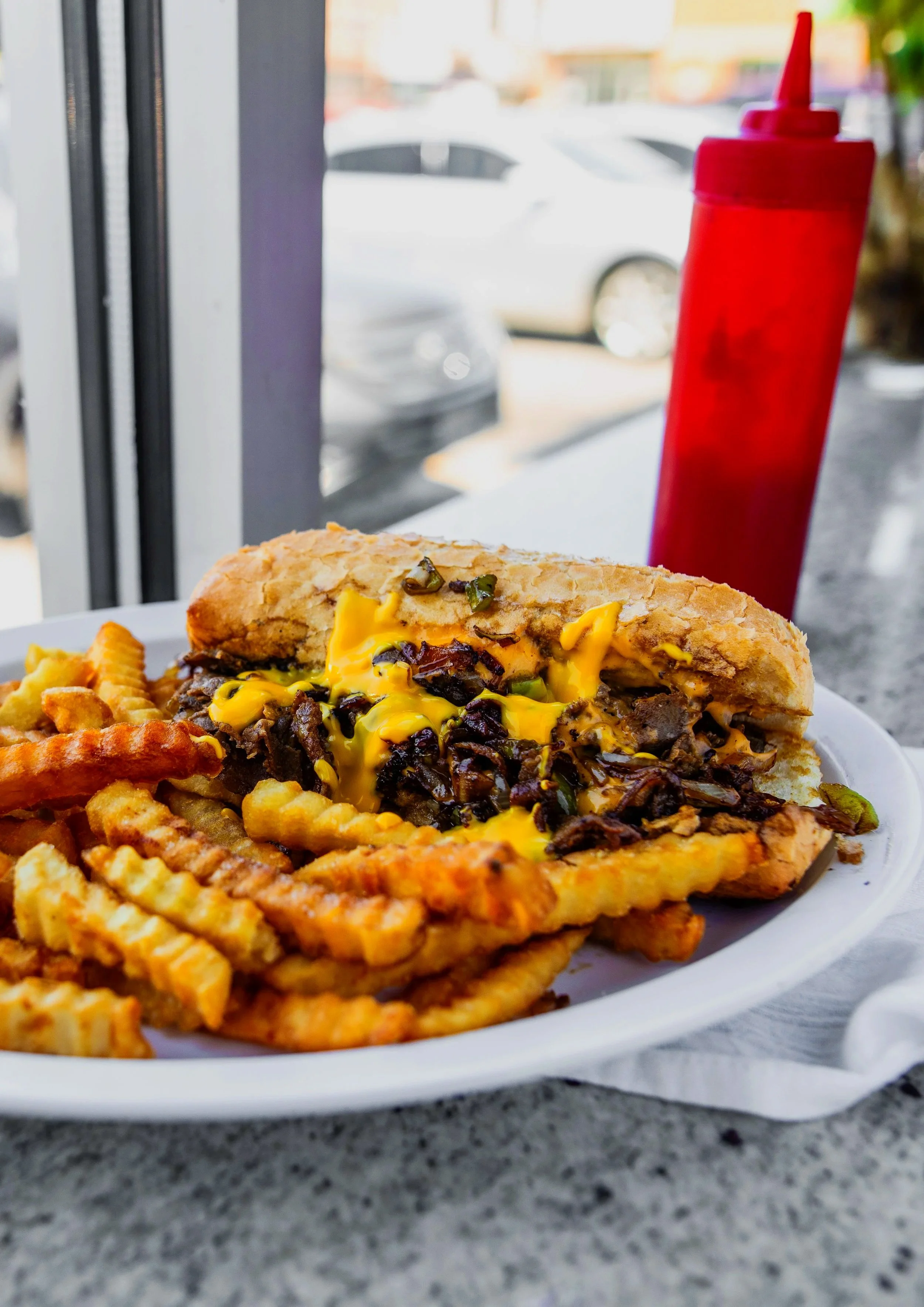 Philly cheesesteak sandwich with fries on a white plate, ketchup bottle in the background, at a restaurant or diner.