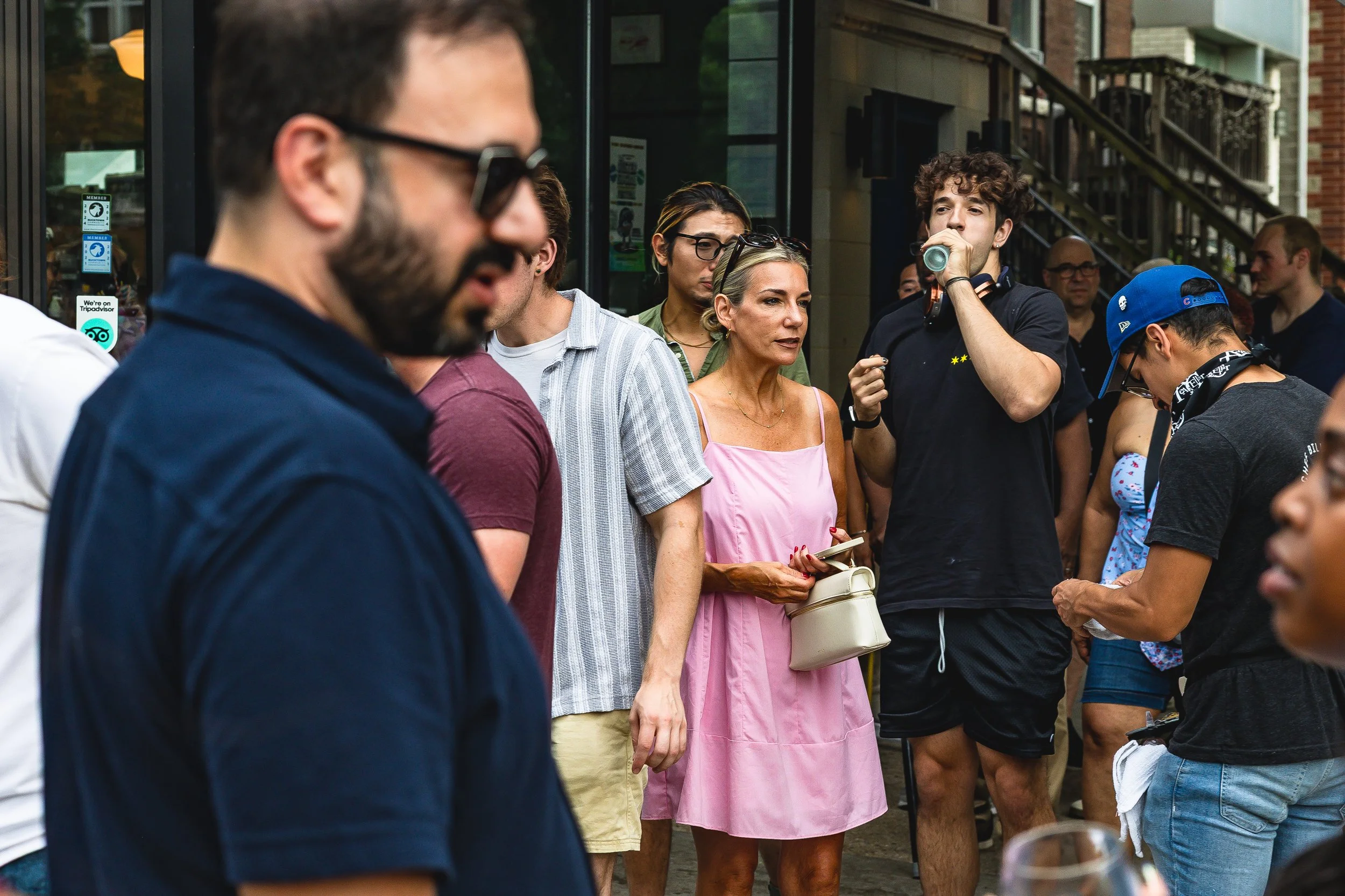 A group of people standing outside a building, some drinking, some looking down, and others looking around. The group includes men and women of diverse ages, including a woman in a pink dress holding a small purse and a young man drinking from a can.