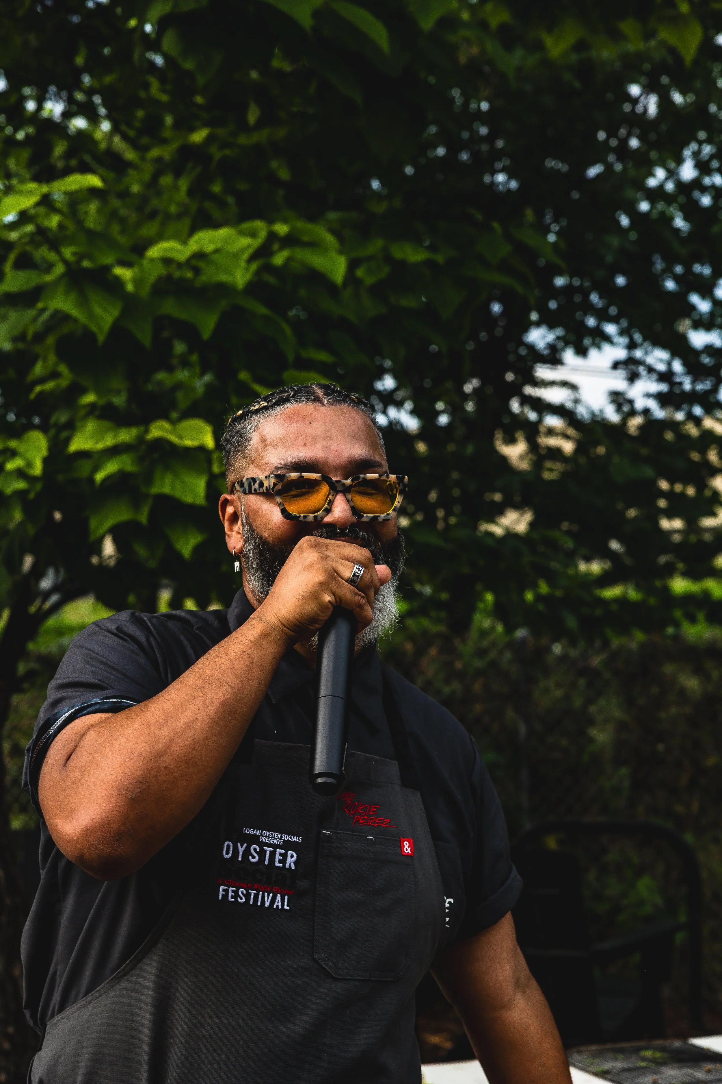 A man with glasses and a beard, wearing a black shirt and apron, speaking into a microphone outdoors under lush green trees.