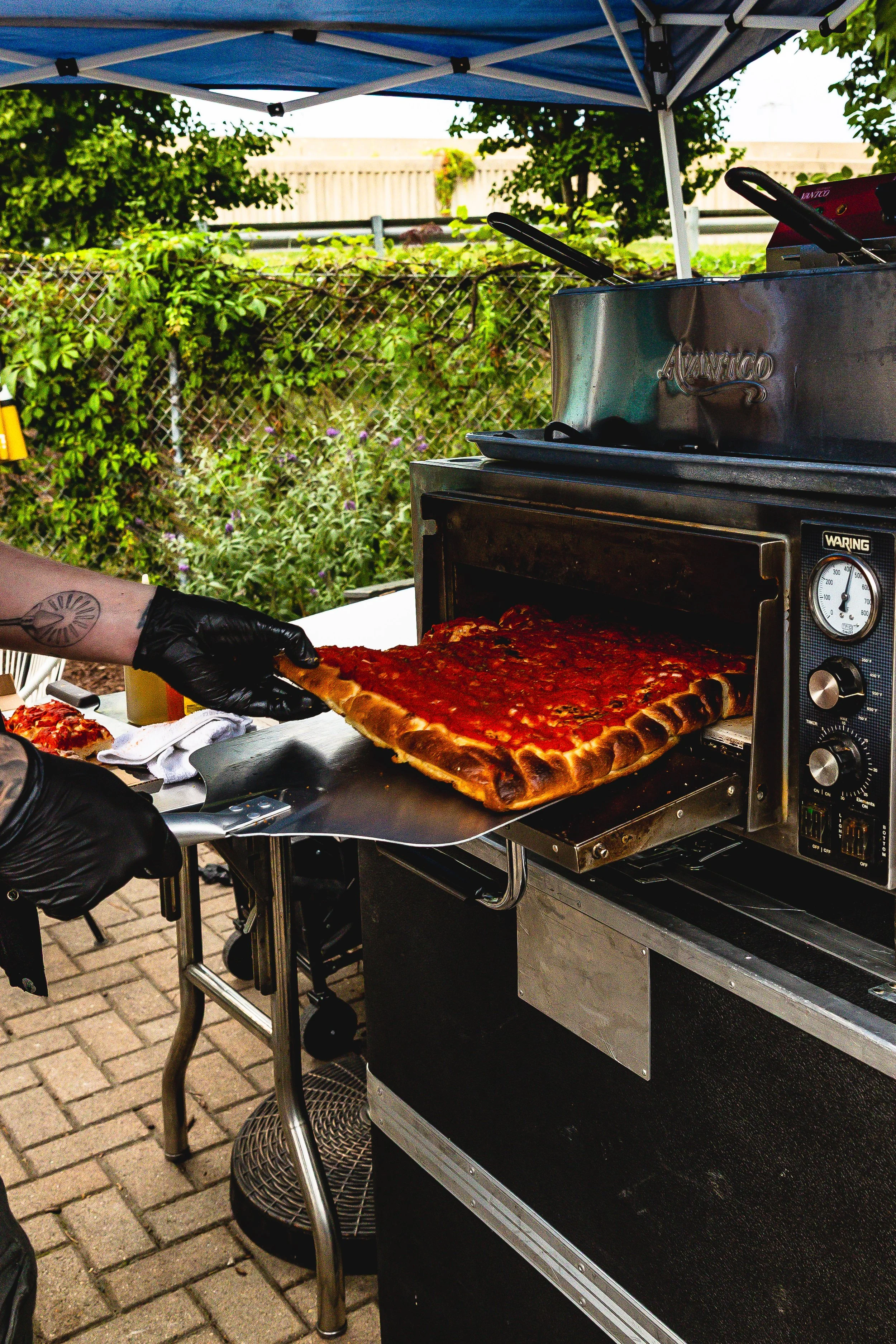 Person wearing black gloves inserting a pizza from a pizza oven outdoors under a blue canopy, with green bushes and a fence in the background.