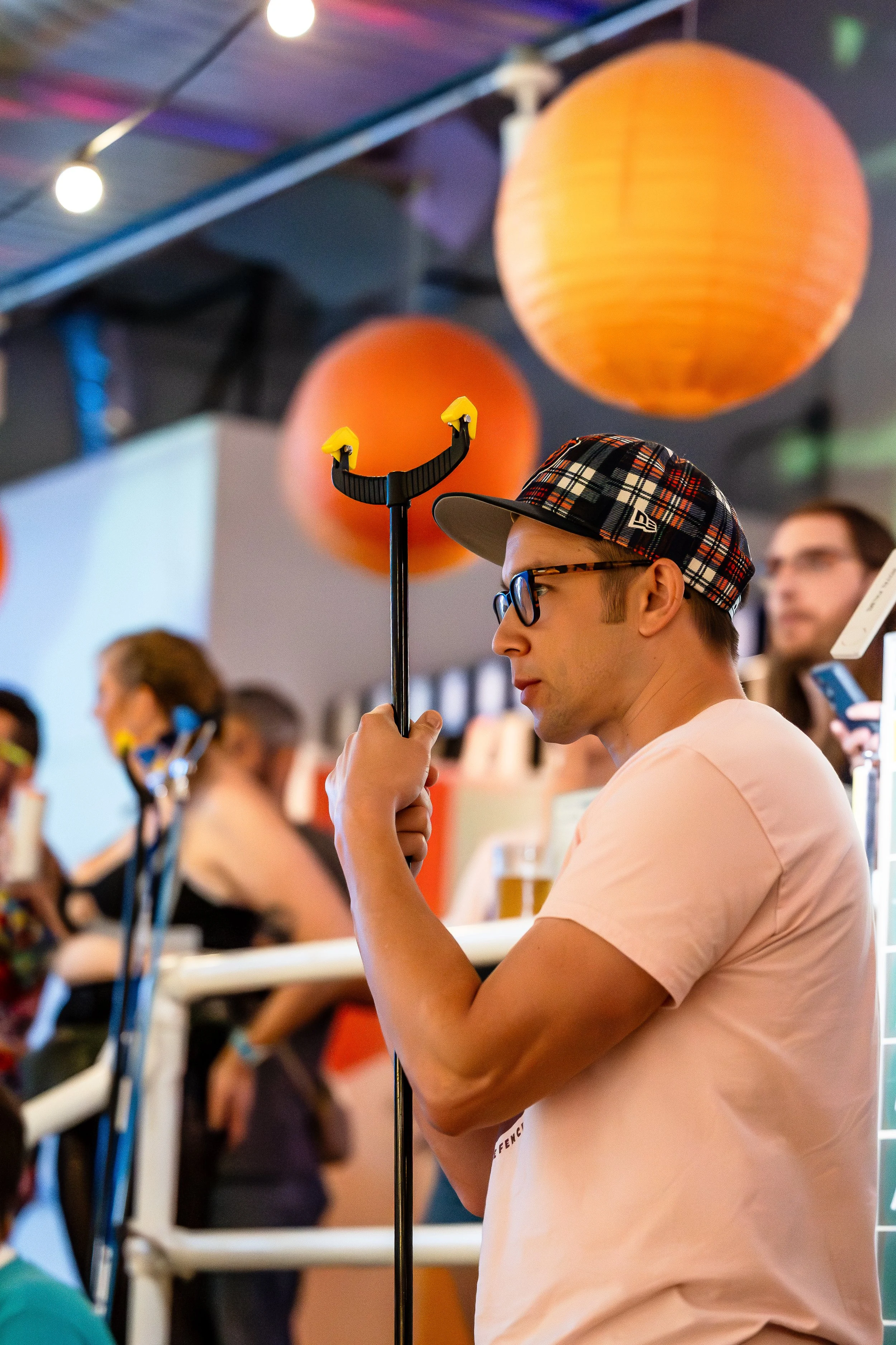 A young man with glasses and a plaid cap is holding a crutch while waiting at a crowded indoor event decorated with orange paper lanterns.
