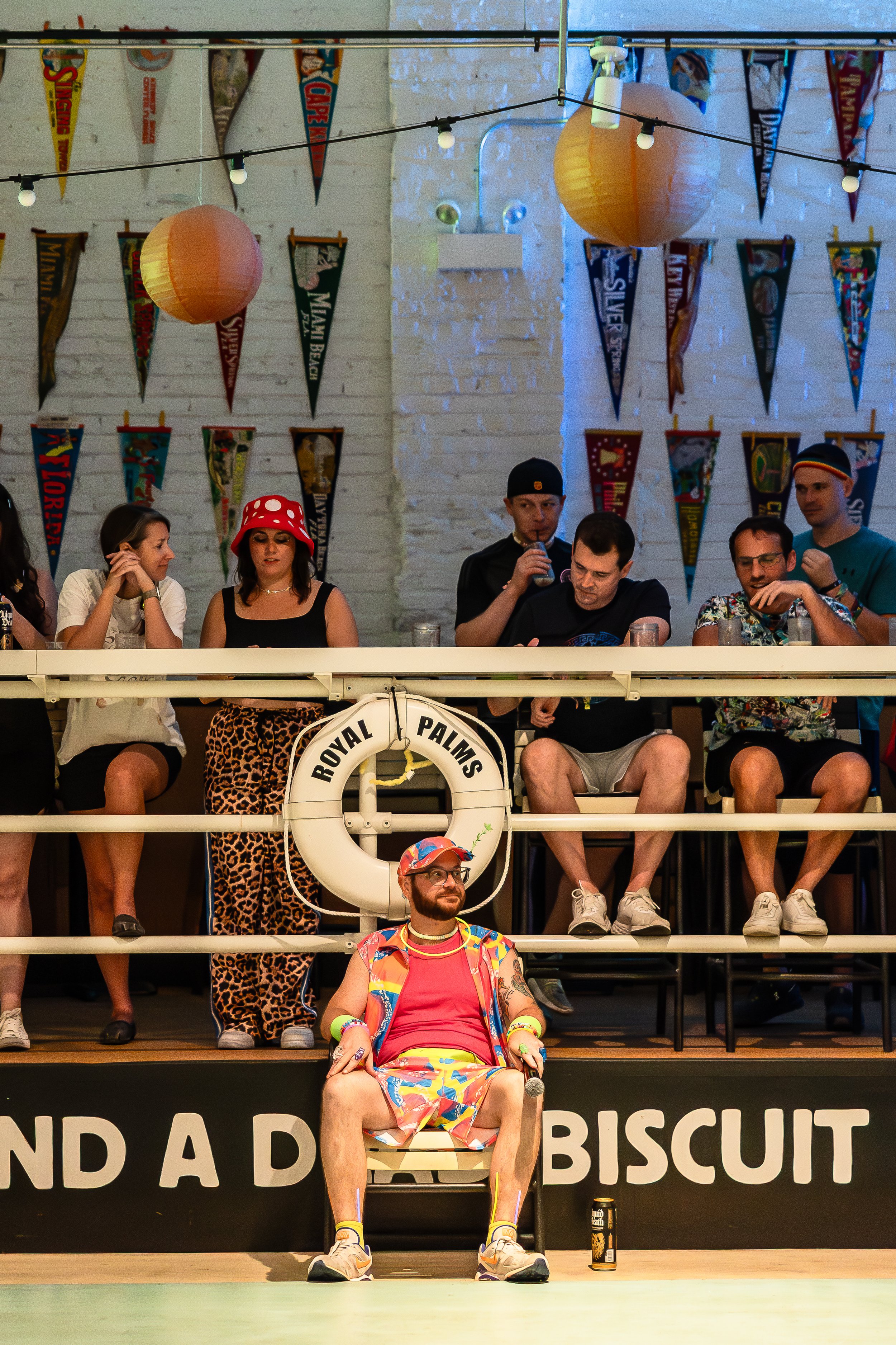 People sitting at a bar with colorful decorations, including pennants and paper lanterns, some wearing summer or costume attire, with a man dressed in colorful summer clothing sitting in front.