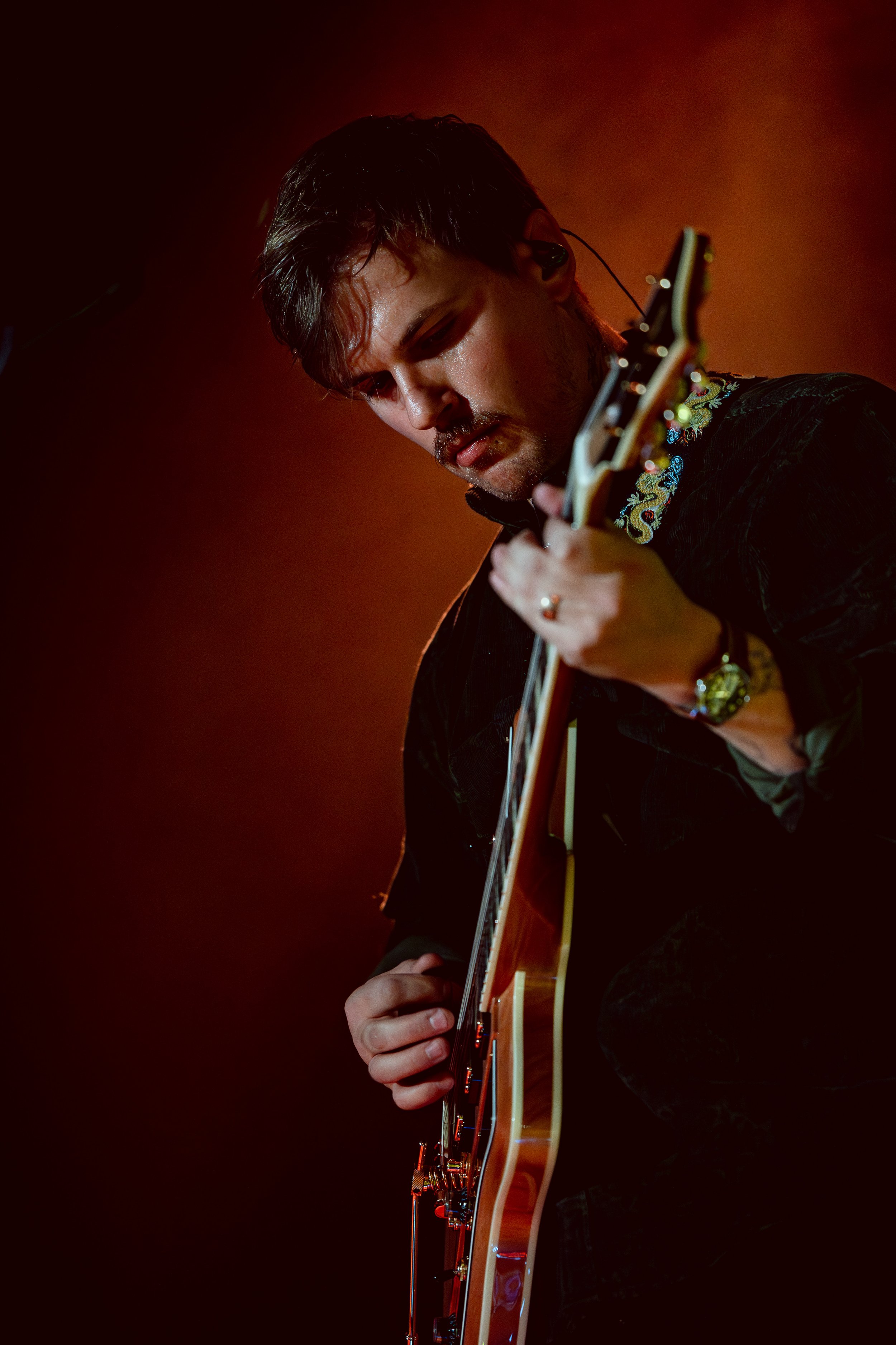 A young man with dark hair playing an electric guitar during a performance, illuminated with warm stage lighting.