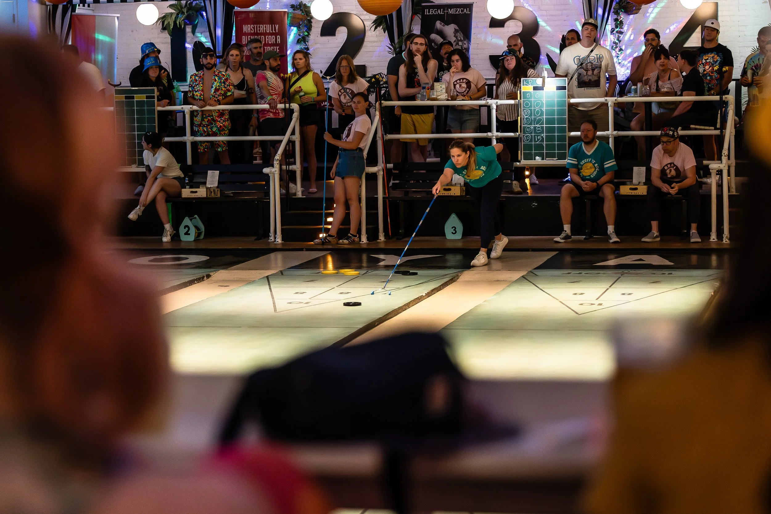 A group of people watching a game of indoor shuffleboard, with one woman actively sliding pucks on the court while others observe from behind a railing.