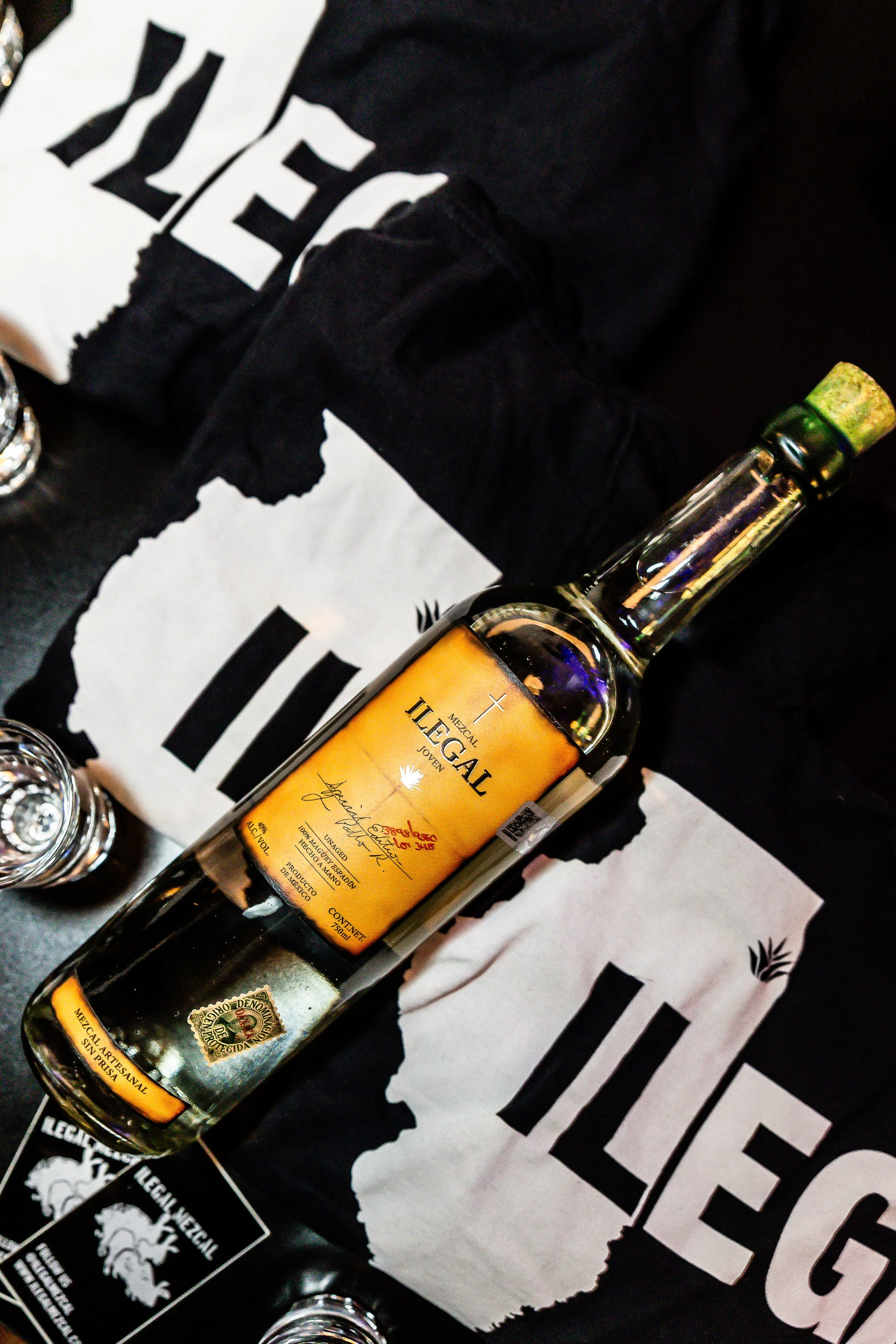 A bottle of Mexican legal aged tequila with a label, lying on a table covered with black and white paper that has the word 'LEGAL' and a torn paper design, surrounded by clear shot glasses.