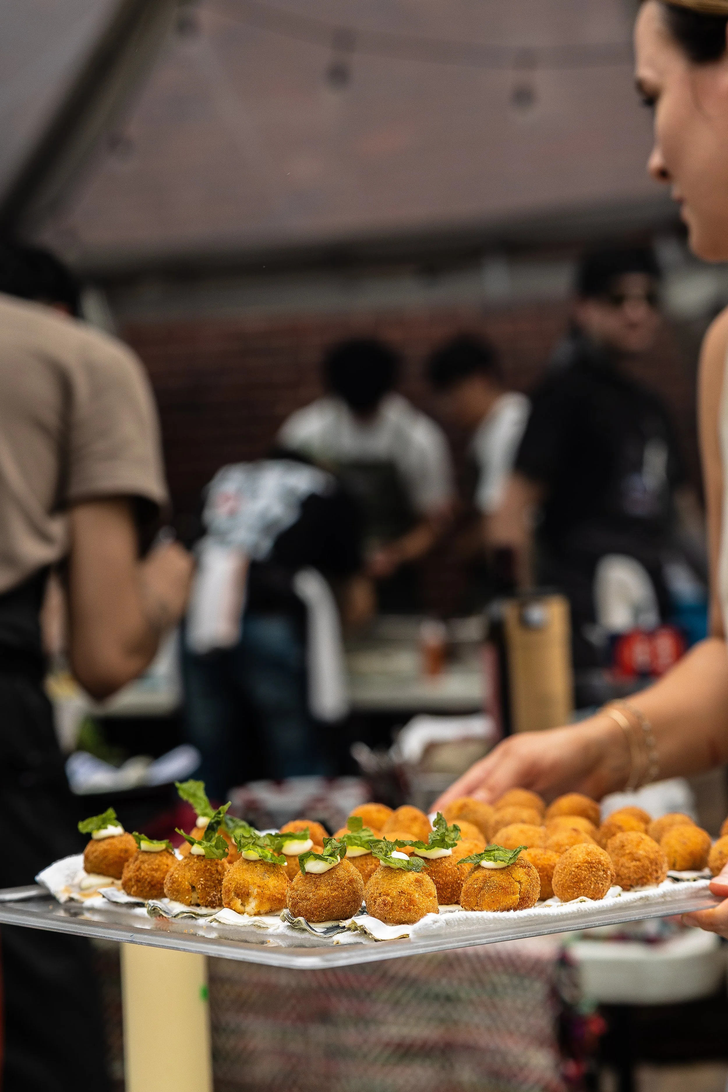 A server holding a tray of fried appetizer balls garnished with herbs, with a busy restaurant kitchen and staff blurred in the background.