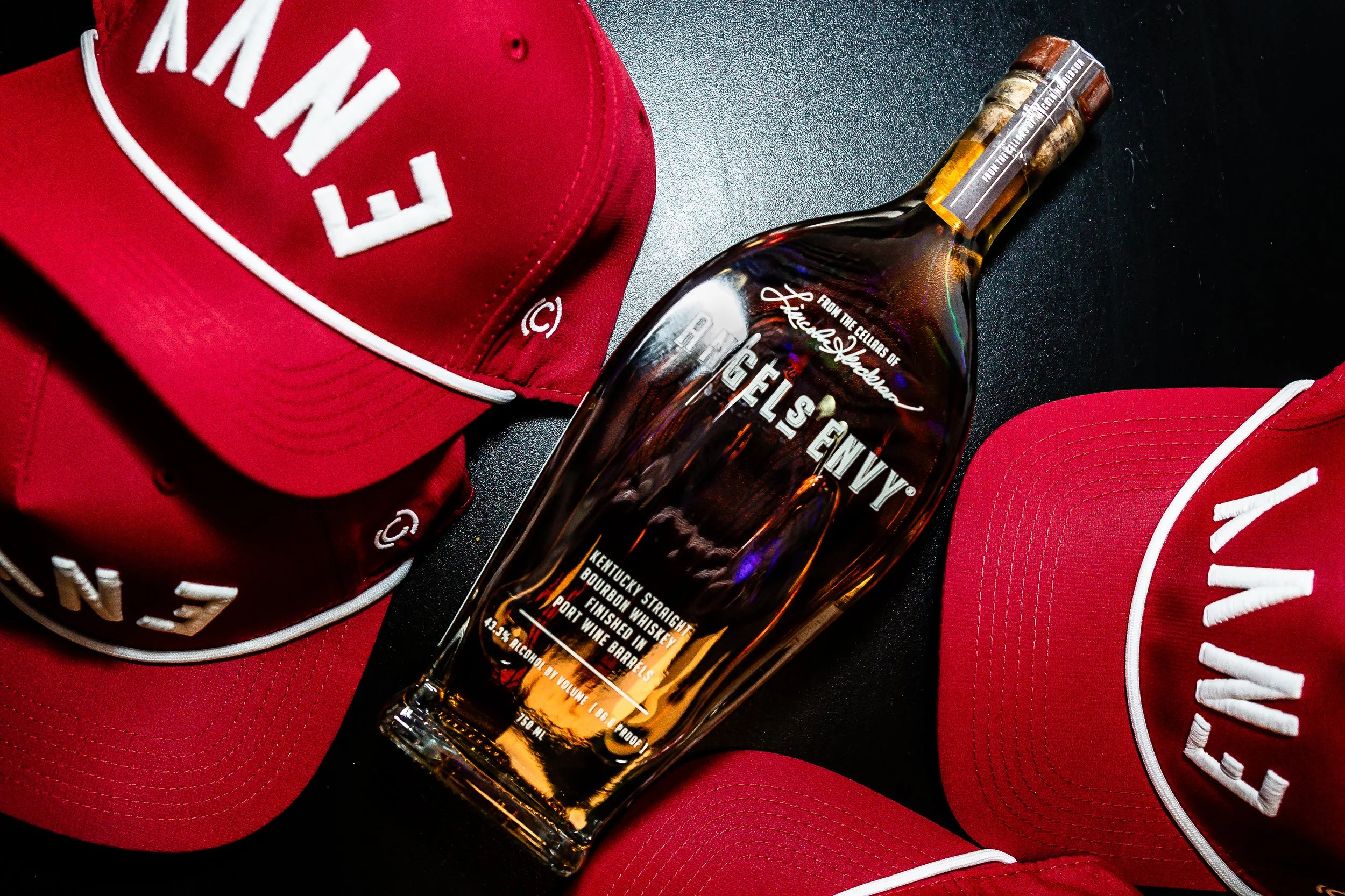 A bottle of Angels Envy bourbon surrounded by three red New Waverly baseball caps with white embroidery on a black surface.