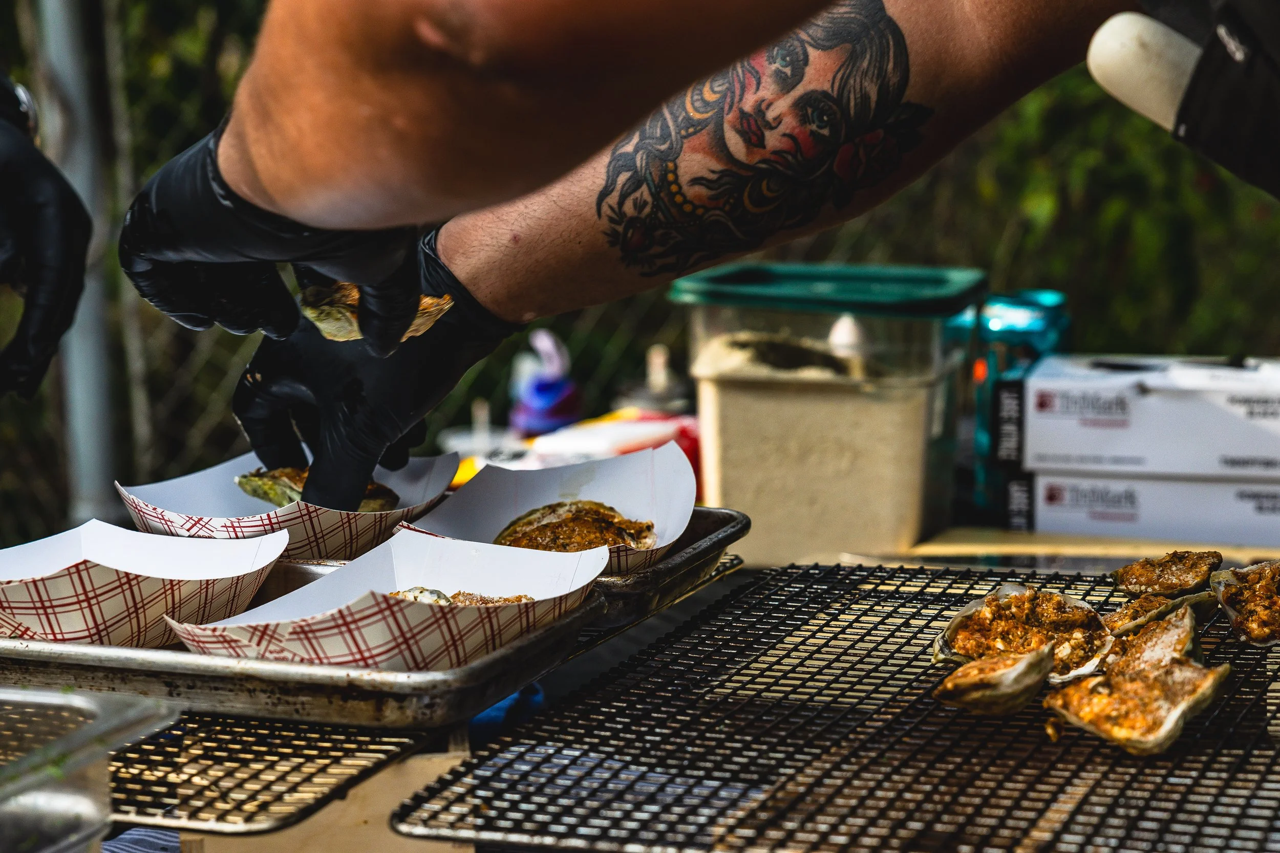 Person wearing black gloves and displaying tattoos on arm preparing food outdoors with several food items, including oysters on a grill and in paper trays, in a setting with containers and packaging in the background.