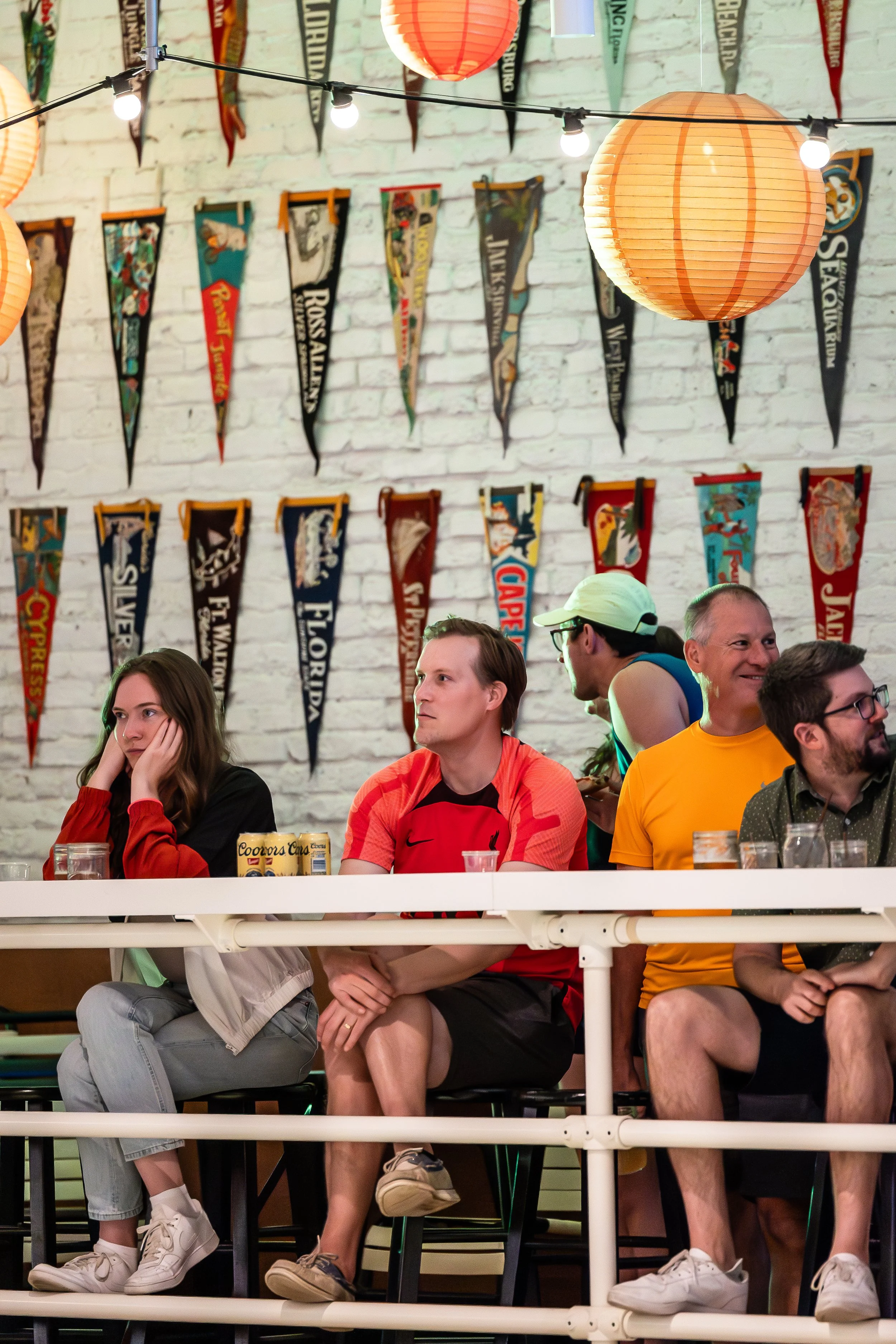 People sitting at a bar with sports pennants hanging on the wall behind them, decorated with paper lanterns and string lights.