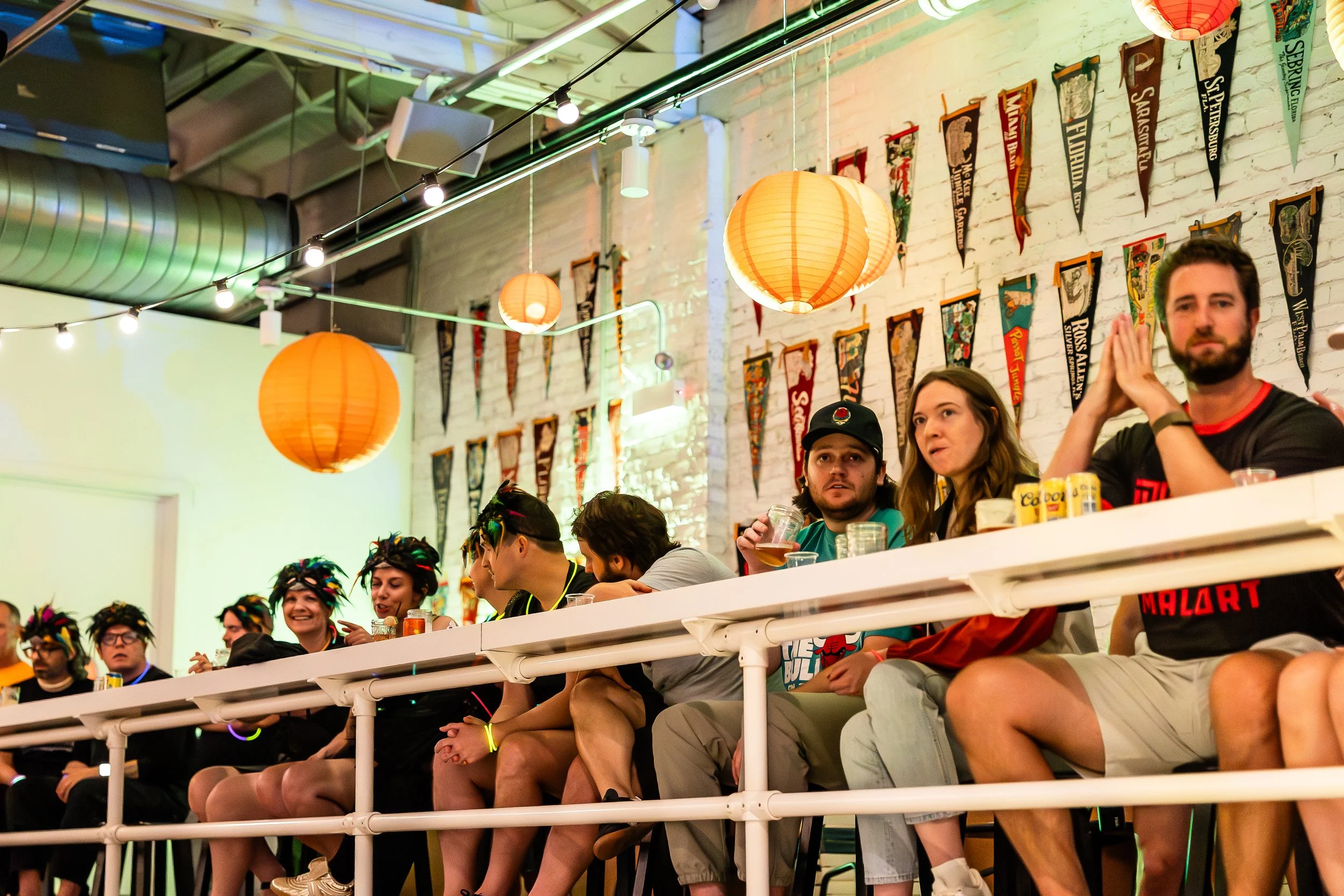 Group of people sitting at a long table in a decorated indoor space with hanging orange lanterns and wall banners, some wearing colorful wigs, appearing to watch or participate in an event.