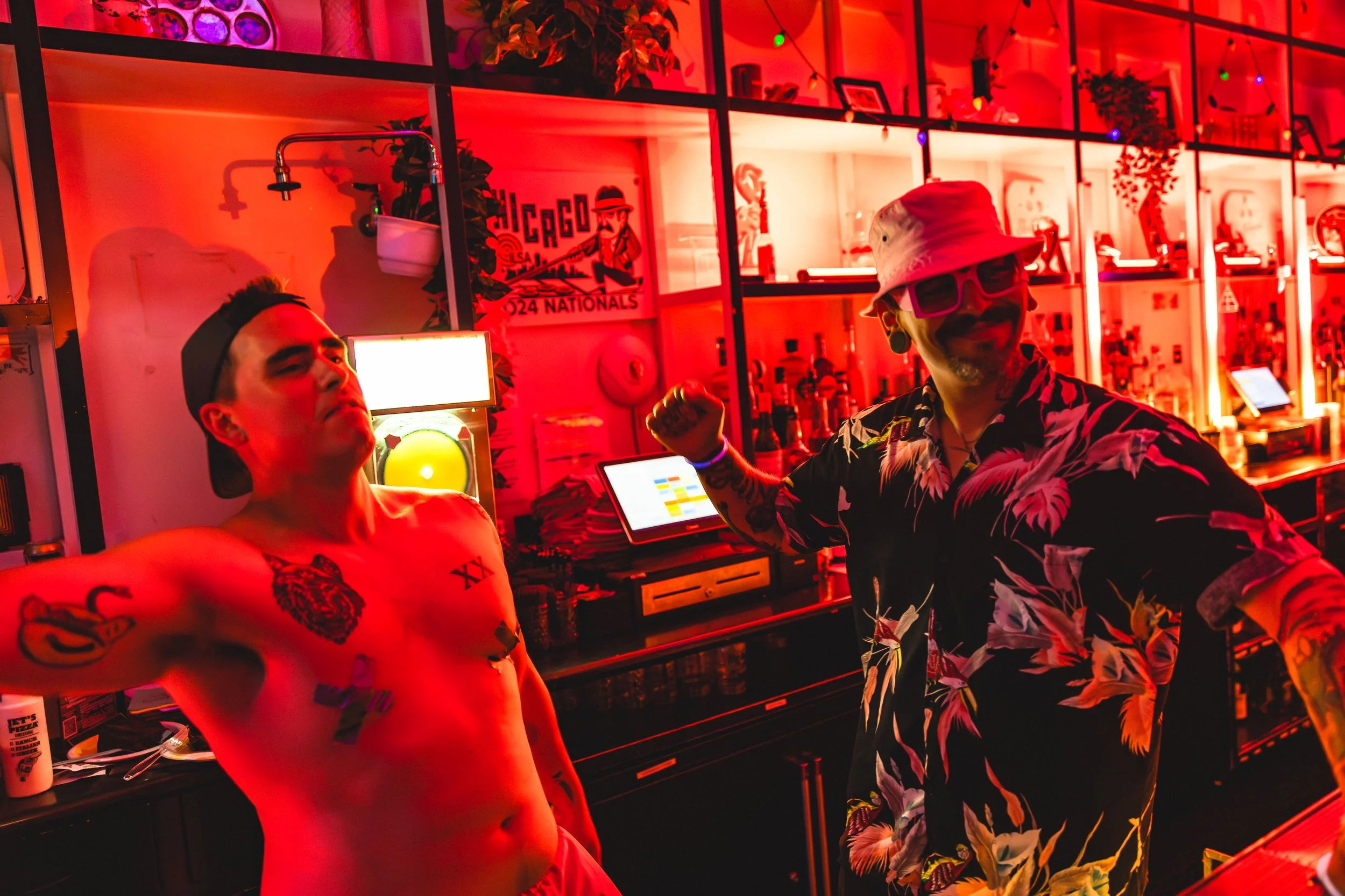 Two men in a vibrant bar with red lighting. One shirtless, tattooed man with a black backward cap making a fist, the other wearing a floral shirt, bucket hat, and sunglasses, smiling. Bar shelves with bottles and decorations in the background.