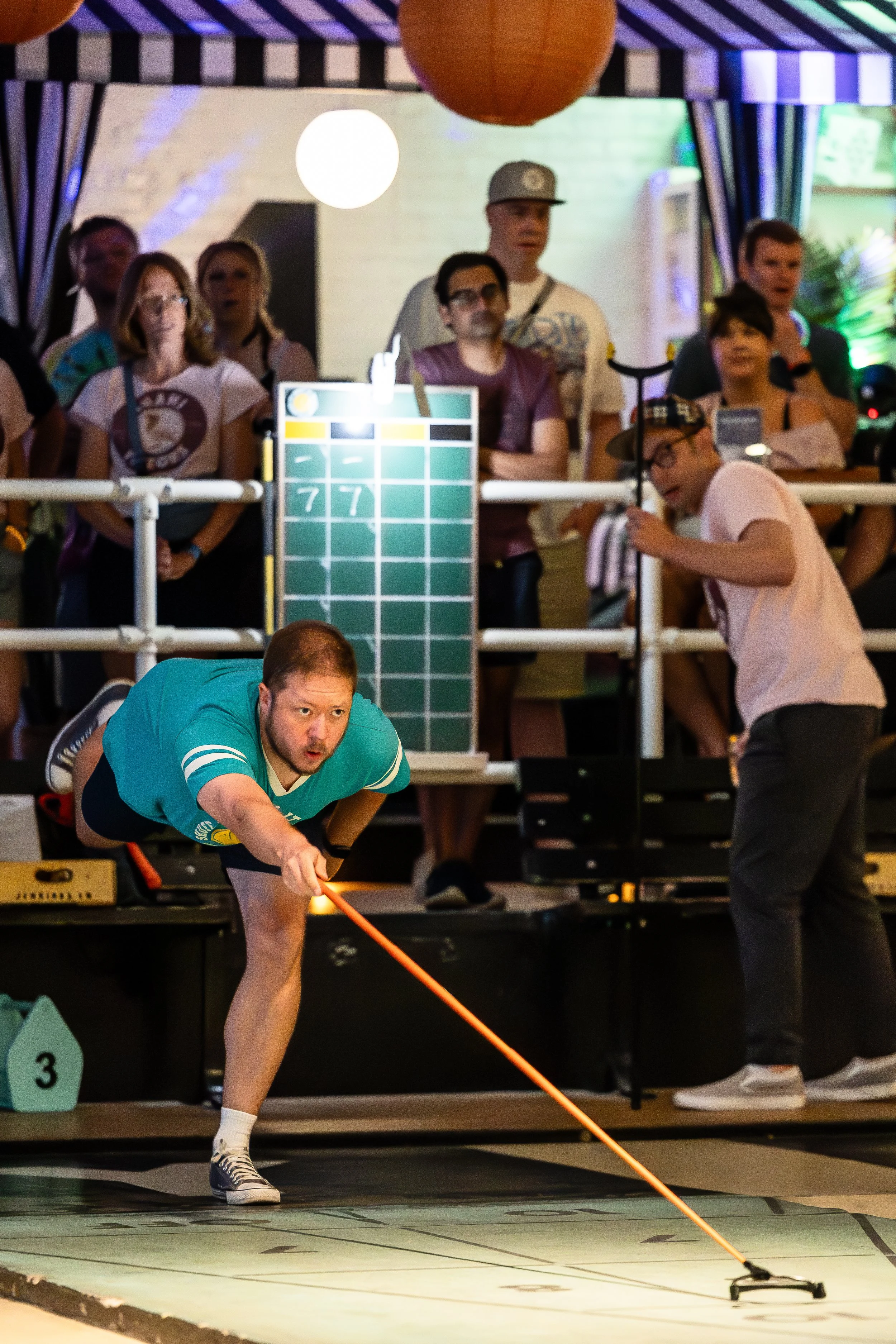 A man is playing shuffleboard, leaning forward and aiming his cue stick at the game surface, with a group of people watching behind him at an indoor event.