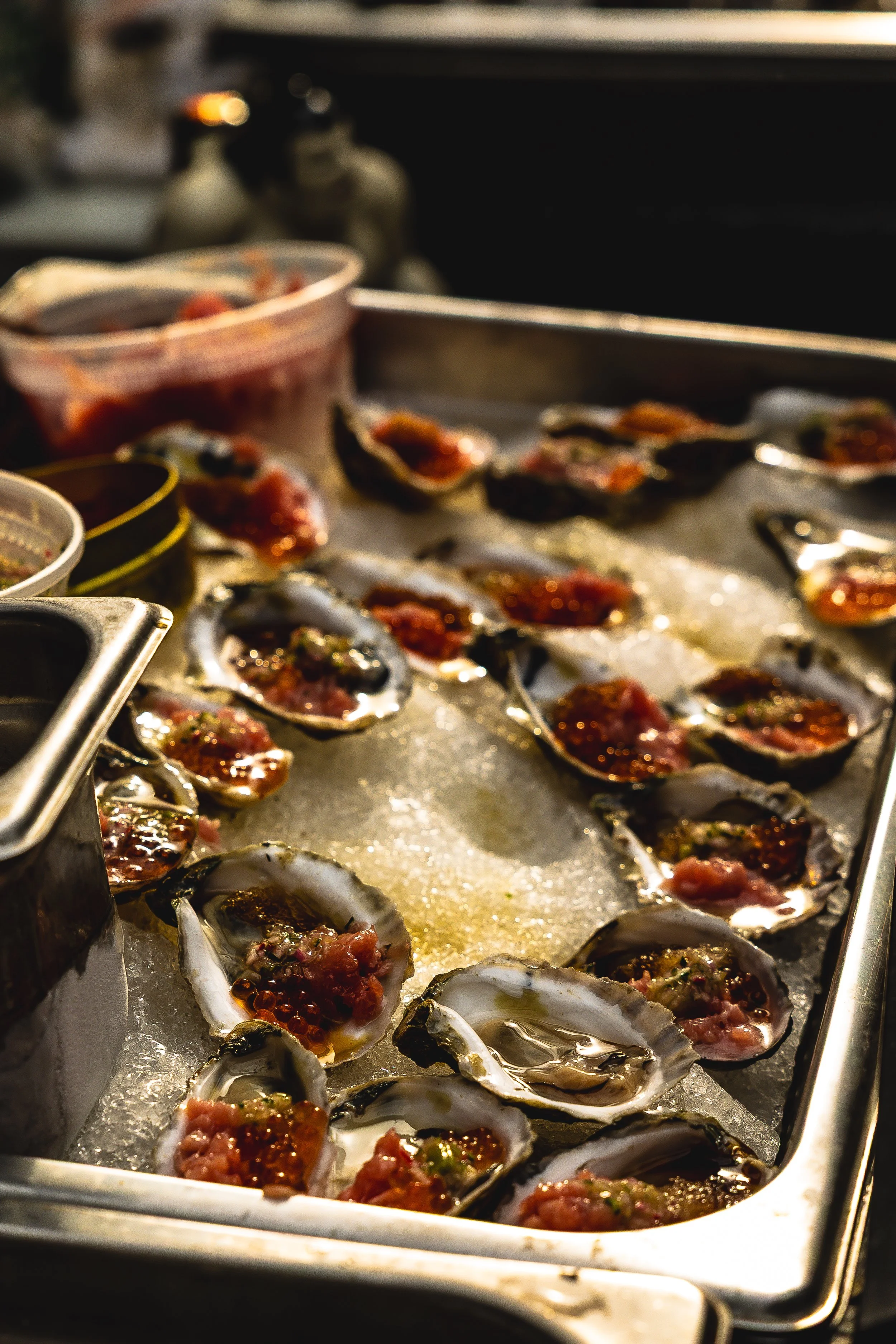 Close-up of oysters on a bed of ice, topped with a sauce or garnish, on a metal tray.