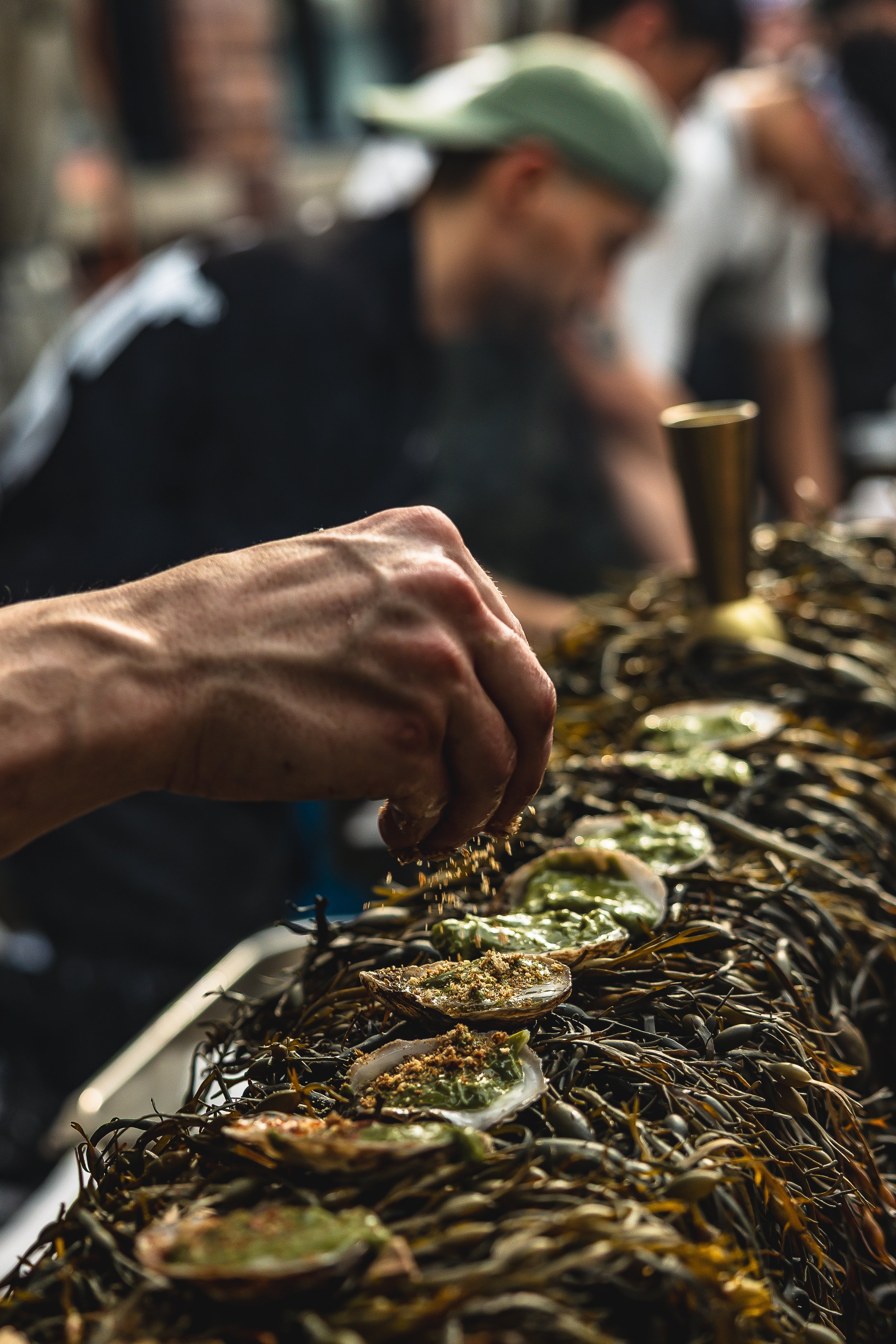 Close-up of a person sprinkling seasoning onto oysters on a bed of seaweed, with several people in the background in a market or food stall setting.