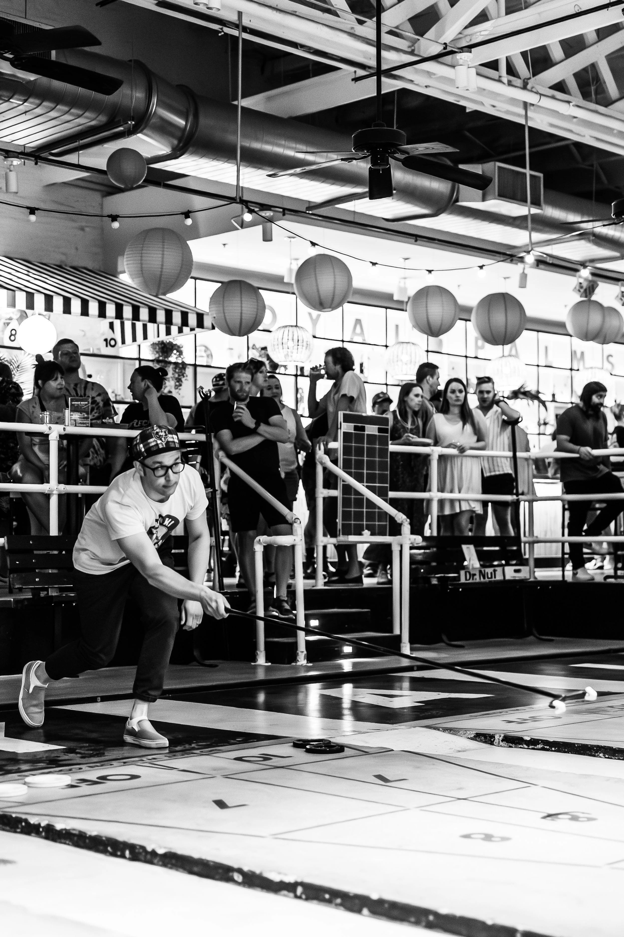 A black and white photo of a man in glasses and a plaid hat playing shuffleboard while a crowd of people watch behind a railing, under hanging paper lanterns at an indoor venue.