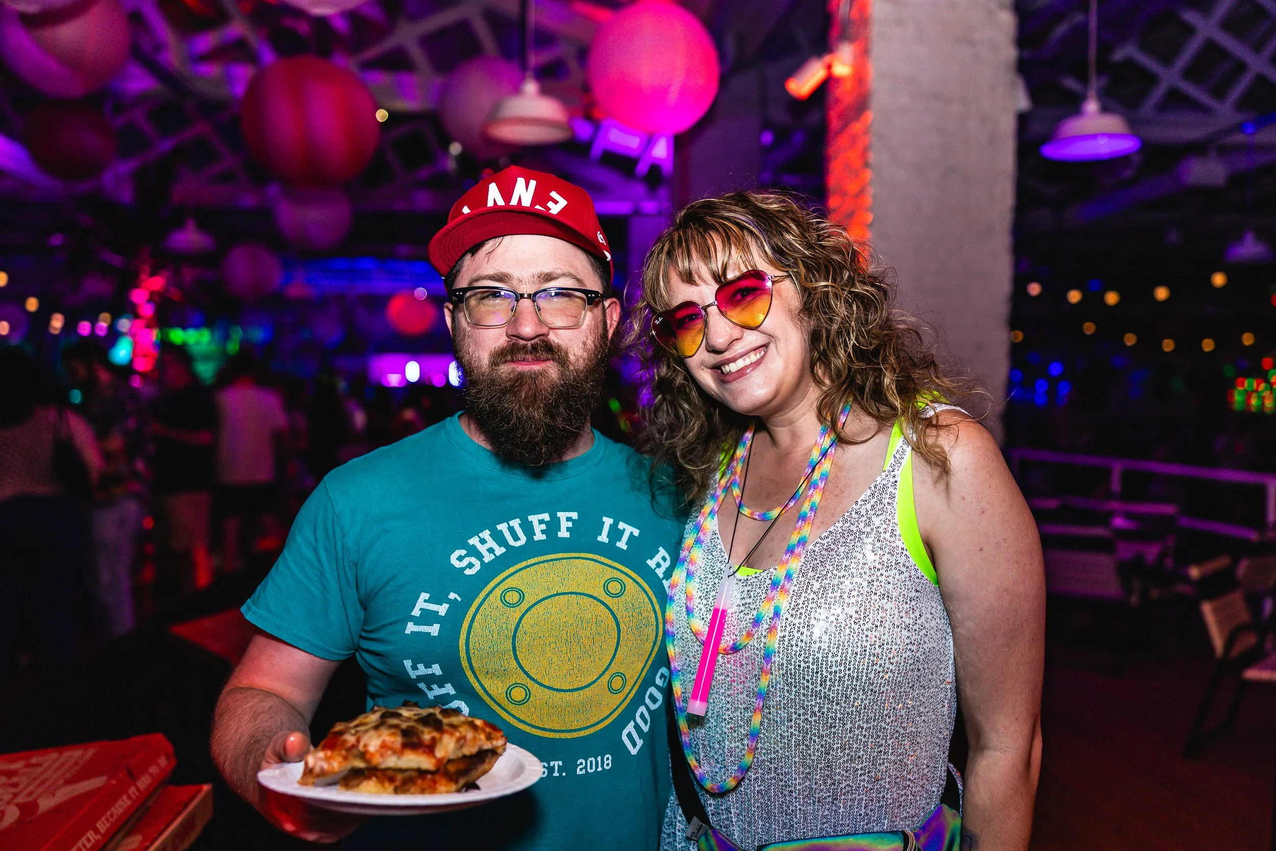 Two people smiling at a party with colorful lighting. The person on the left has a beard, glasses, a red cap, and a blue t-shirt holding a plate with pizza. The person on the right has curly hair, heart-shaped sunglasses, a sequined top, and a rainbo